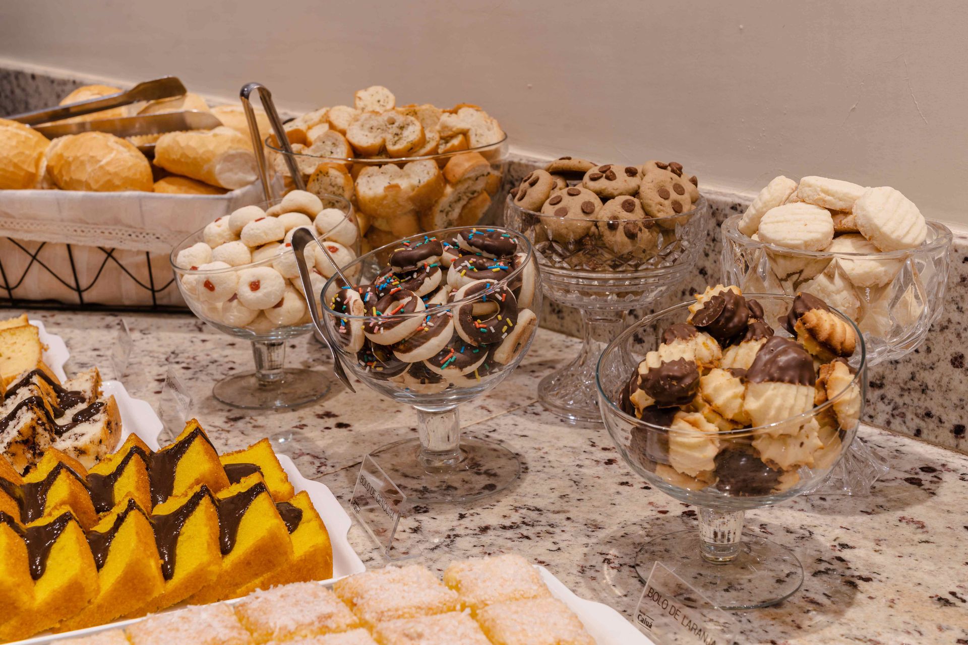 Variedade de doces e biscoitos em recipientes de vidro dispostos em uma mesa de bufê.