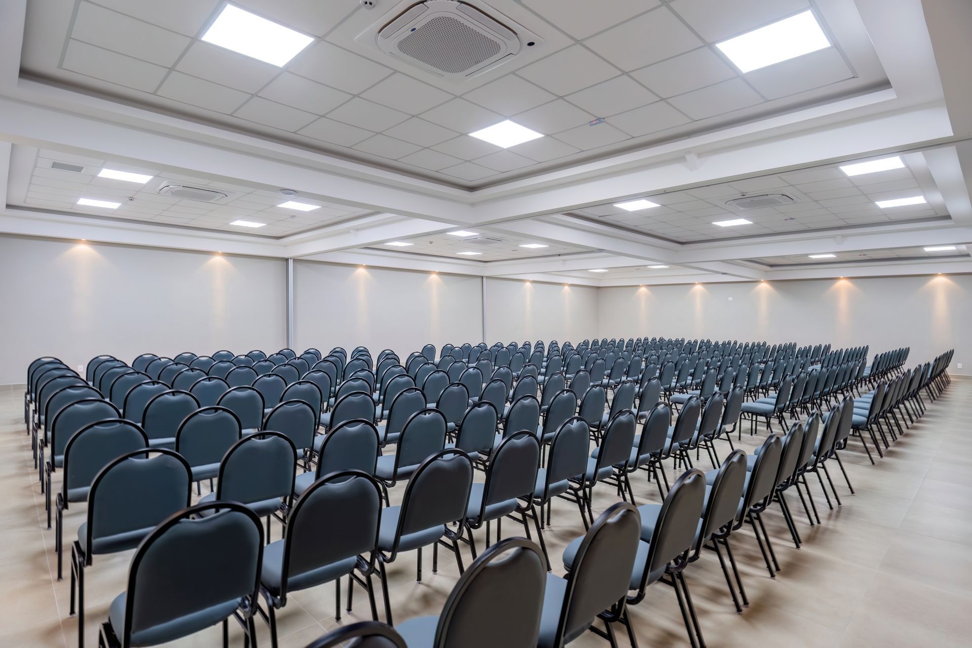 Auditório vazio com fileiras de cadeiras cinzentas, paredes brancas e luzes no teto.
