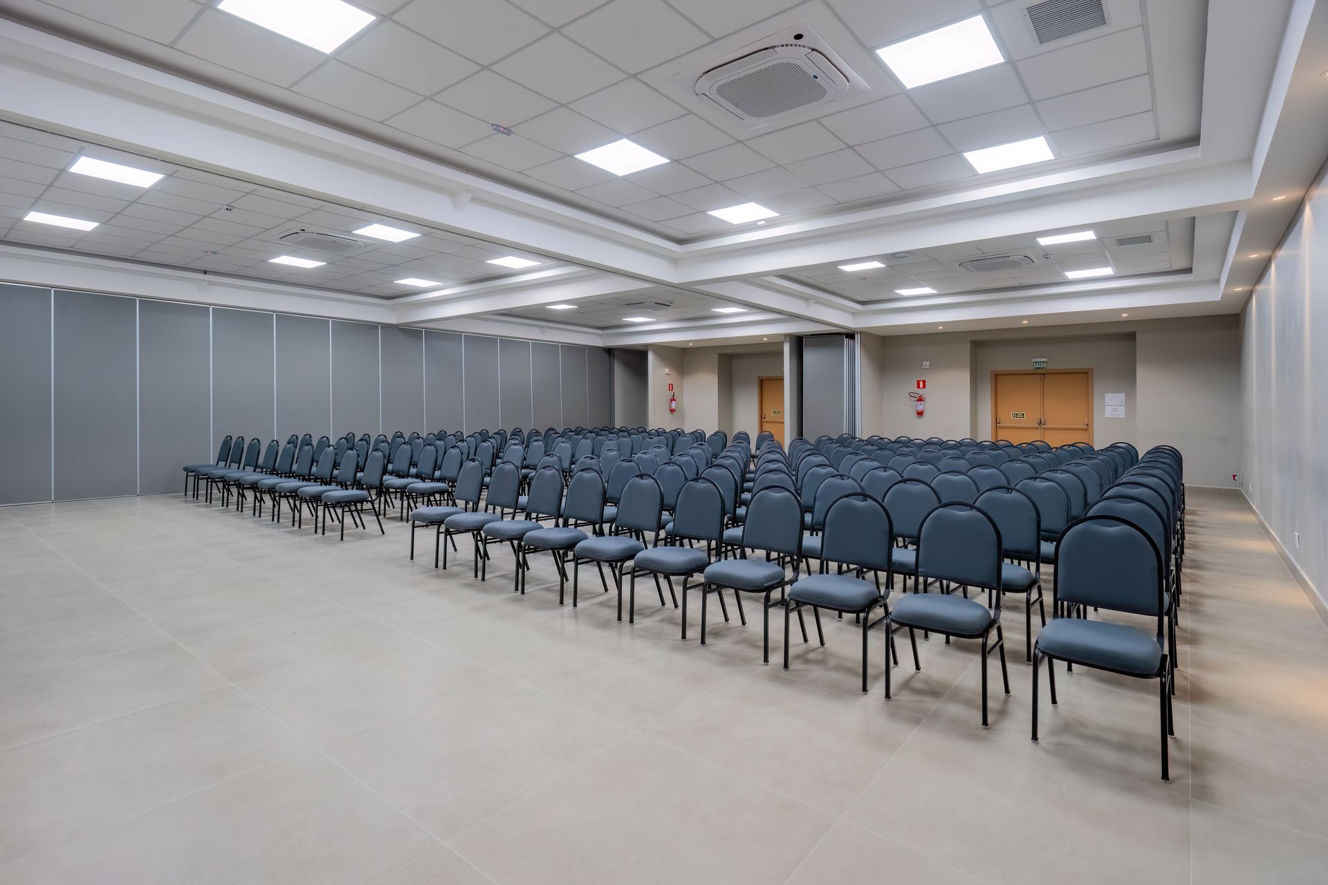 Auditório vazio com fileiras de cadeiras azuis sob luzes brilhantes no teto.