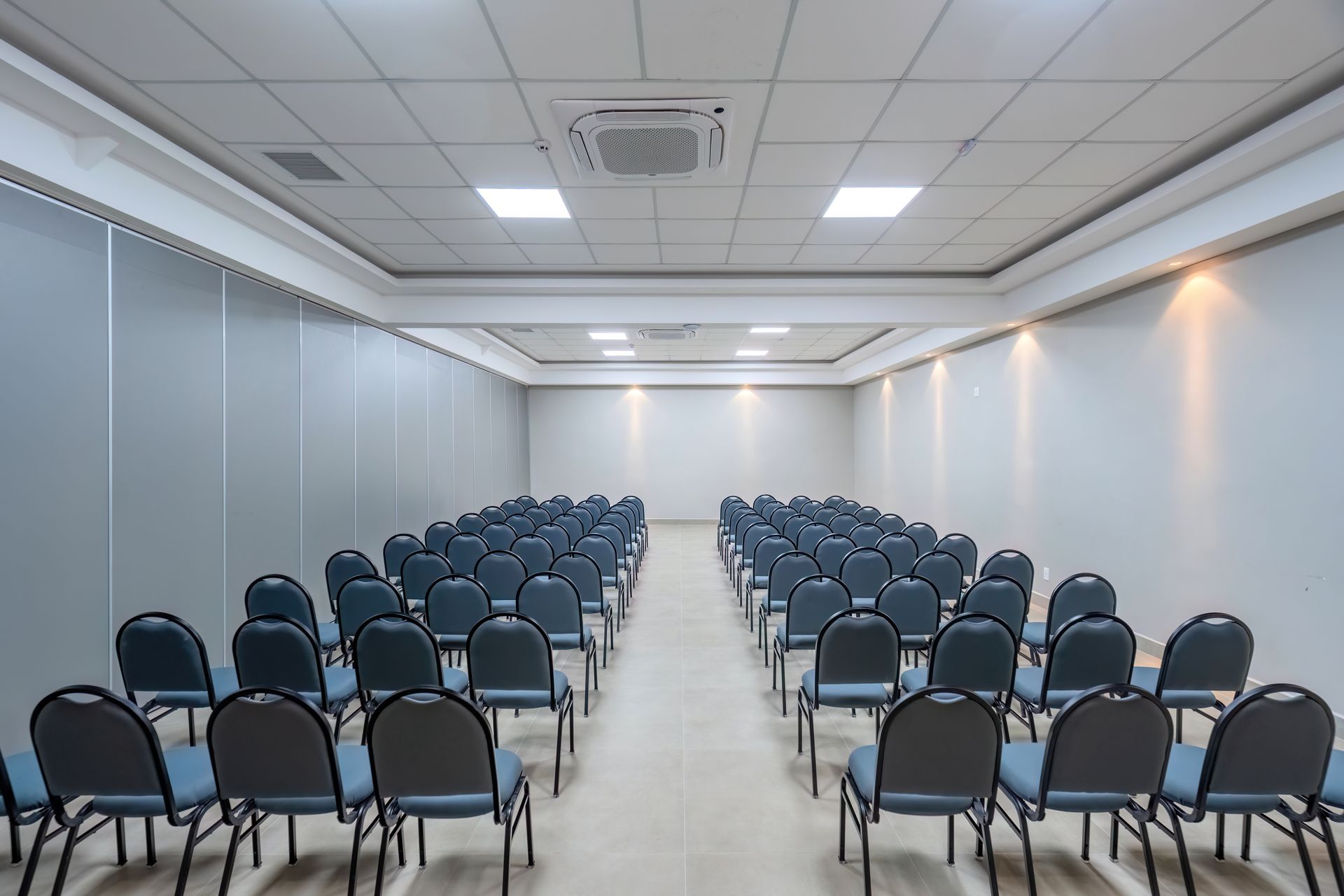 Sala de conferências vazia com fileiras de cadeiras azuis, paredes cinzentas e luzes brilhantes no teto.