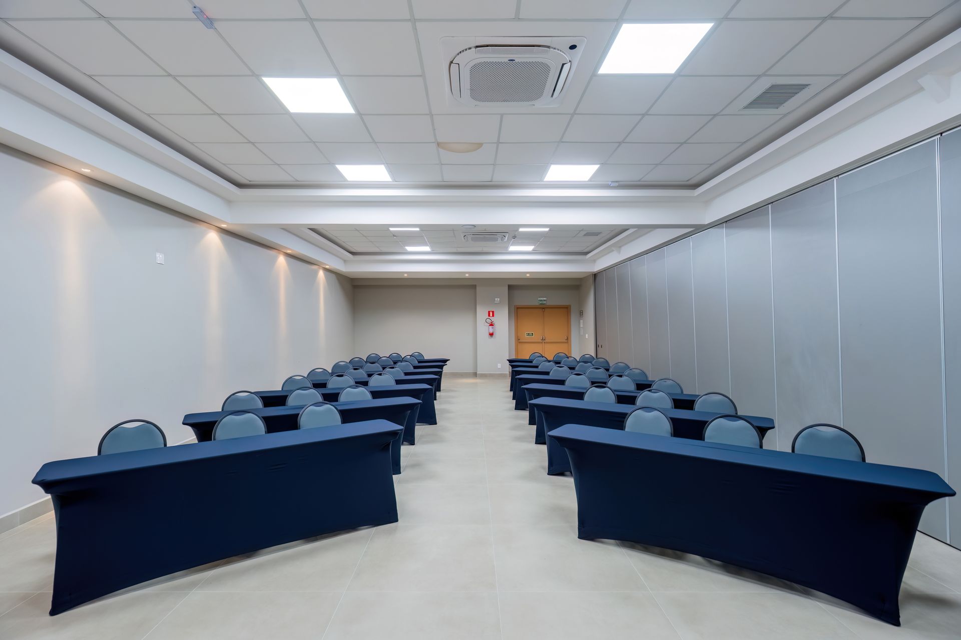 Sala de conferências com fileiras de mesas e cadeiras com capas azuis. Paredes e teto brancos, aparelho de ar condicionado.