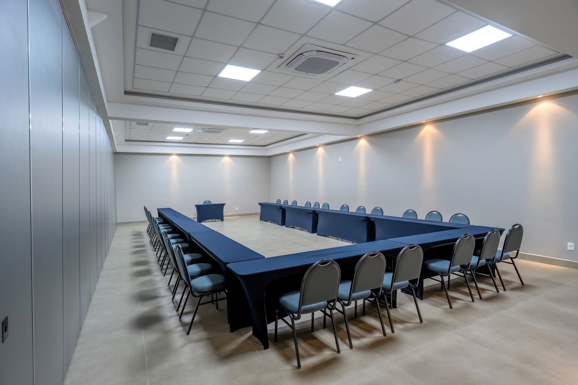 Sala de conferências com mesa e cadeiras em formato de U. Paredes e teto em tons claros.