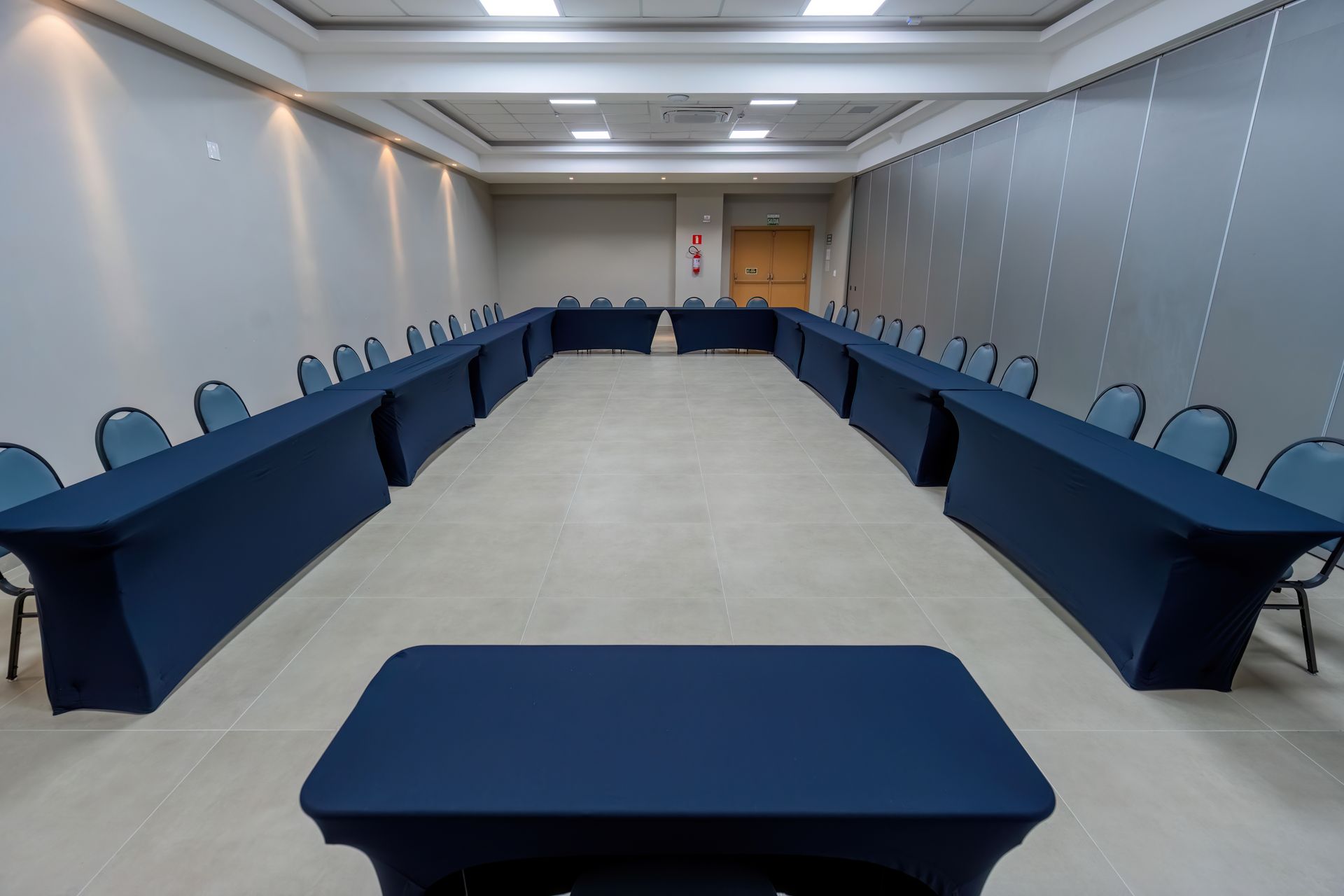 Sala de conferências com mesas e cadeiras azuis dispostas em forma de U.