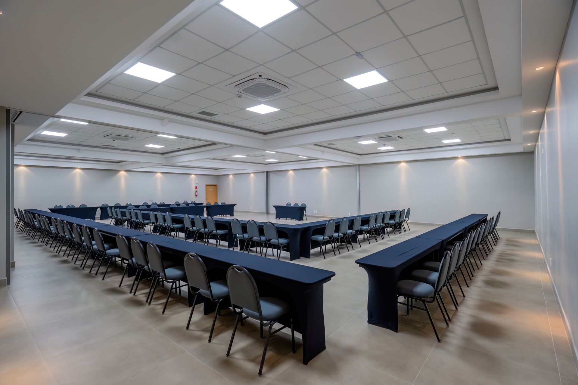 Sala de conferências com mesas em formato de U, toalhas de mesa azul-escuras, cadeiras cinza e iluminação embutida.