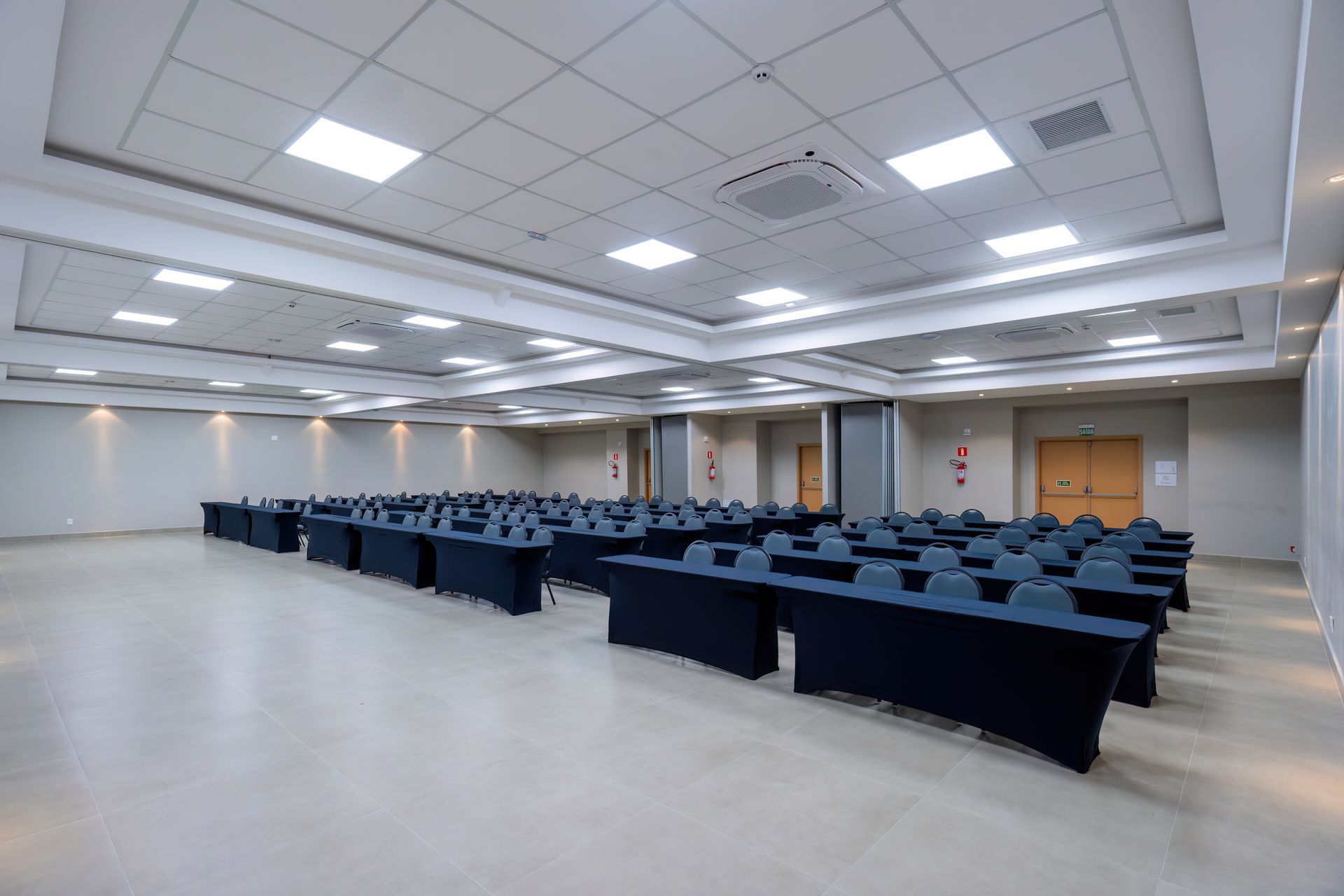 Sala de conferências vazia com fileiras de mesas e cadeiras pretas, paredes e teto brancos.