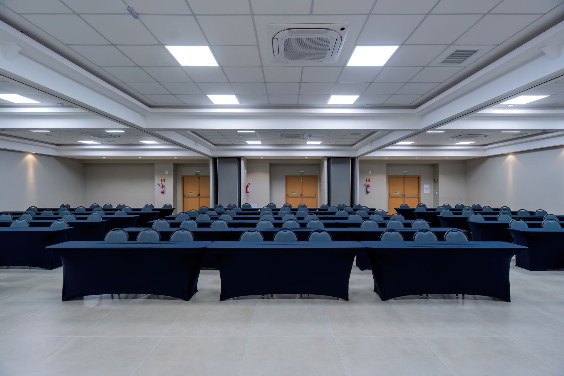 Sala de conferências vazia com fileiras de mesas cobertas com toalhas pretas.