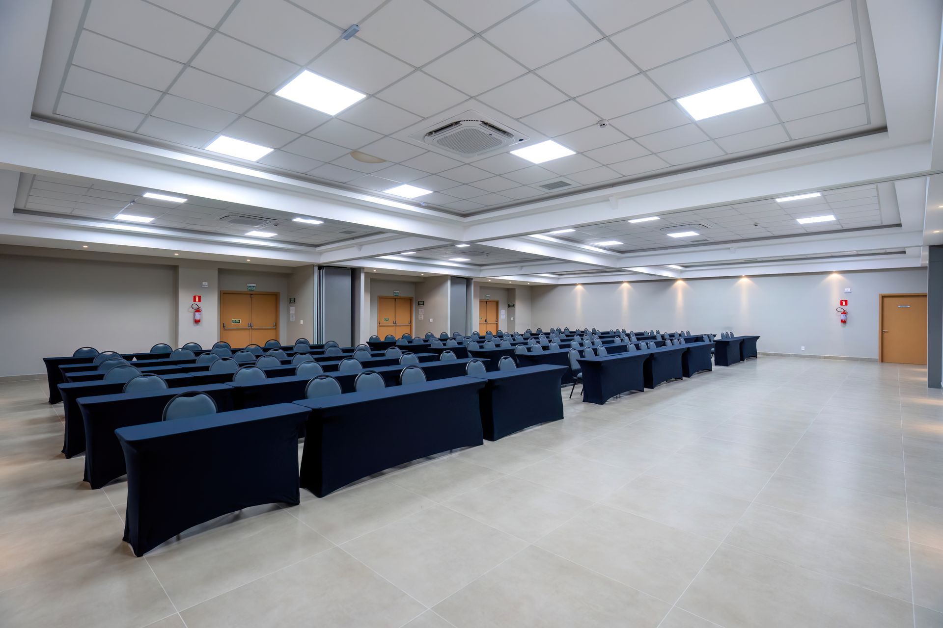 Sala de eventos com fileiras de mesas cobertas com toalhas, cadeiras azuis, paredes em tons neutros e iluminação no teto.