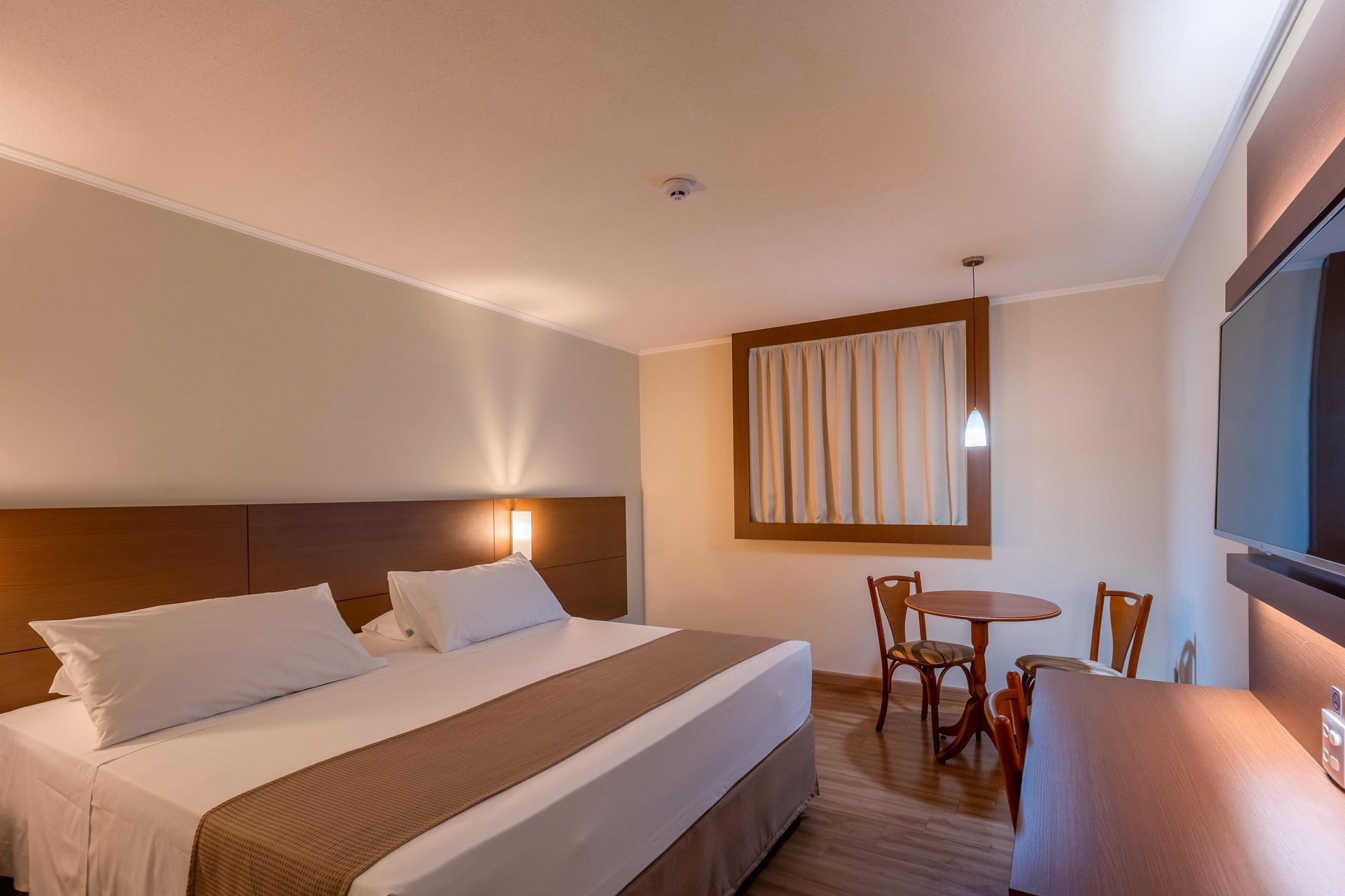 Quarto de hotel com cama, mesa pequena com duas cadeiras e janela.