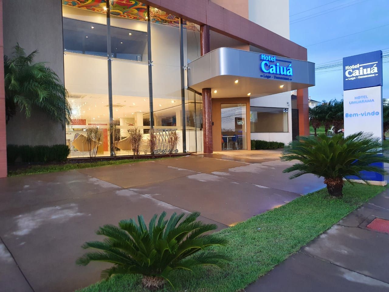 Entrada do hotel ao entardecer, hall de entrada envidraçado, letreiro iluminado e calçada molhada ladeada por plantas tropicais.