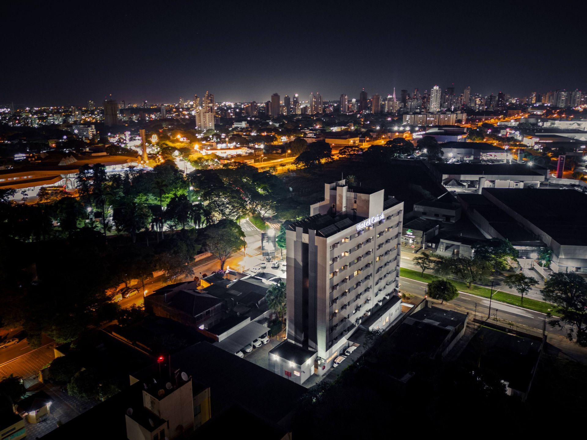 Vista aérea noturna de um prédio alto e iluminado em uma cidade com muitas luzes ao fundo.