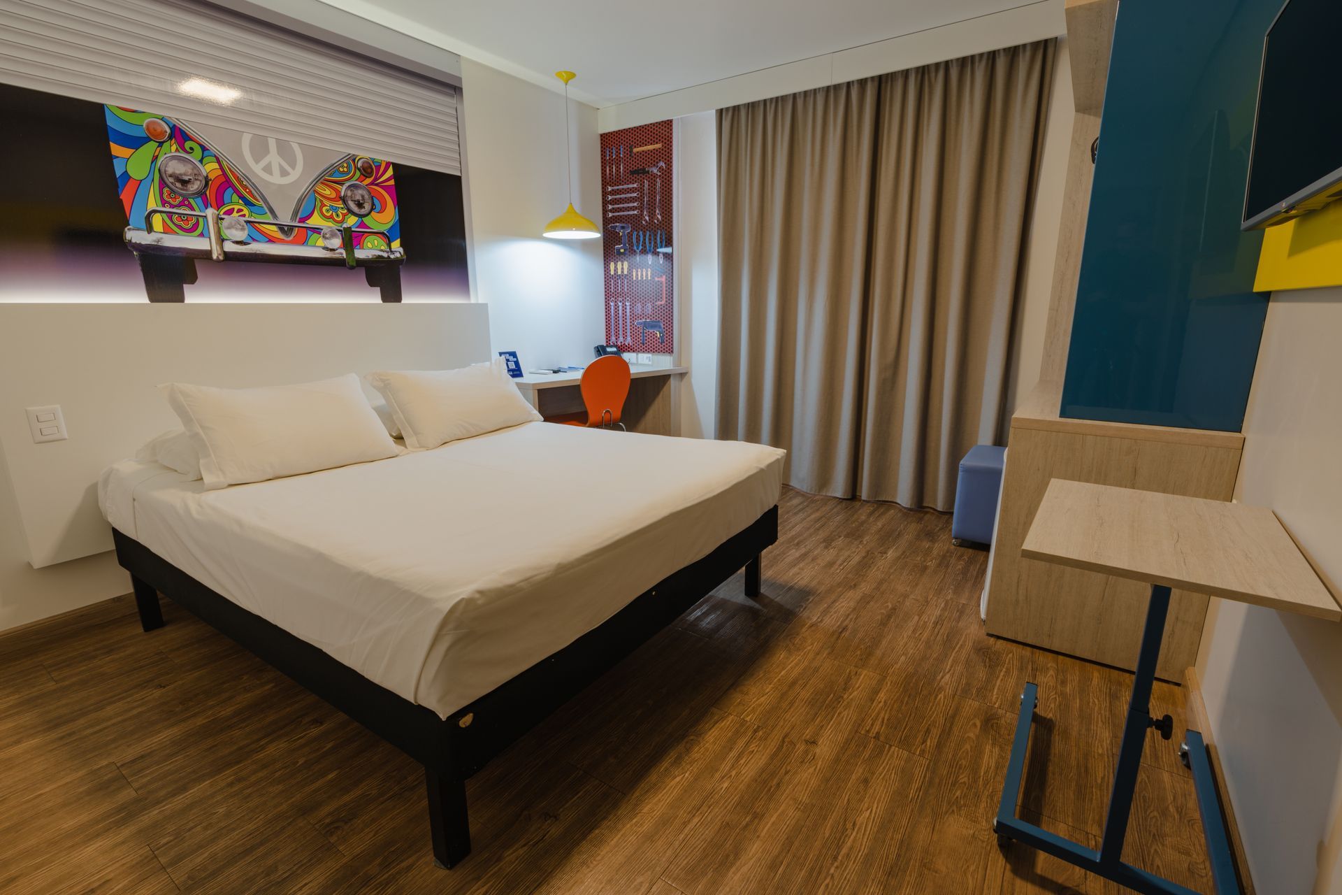 Quarto de hotel com cama, escrivaninha com TV, cadeira e sofá preto. Cortinas bege e piso de madeira.