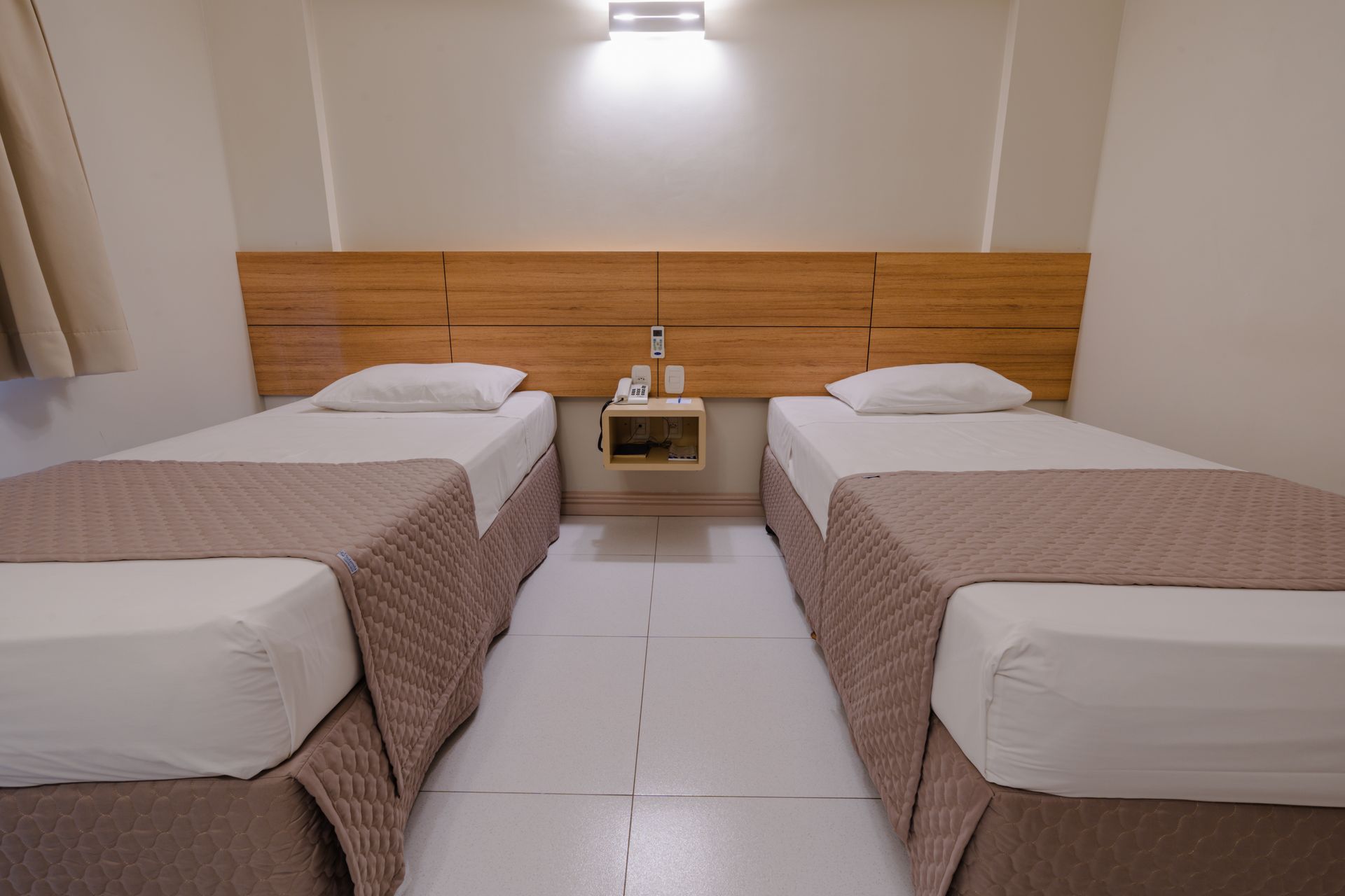 Quarto de hotel com duas camas de solteiro, escrivaninha e TV. Decoração em tons de marrom claro e bege.
