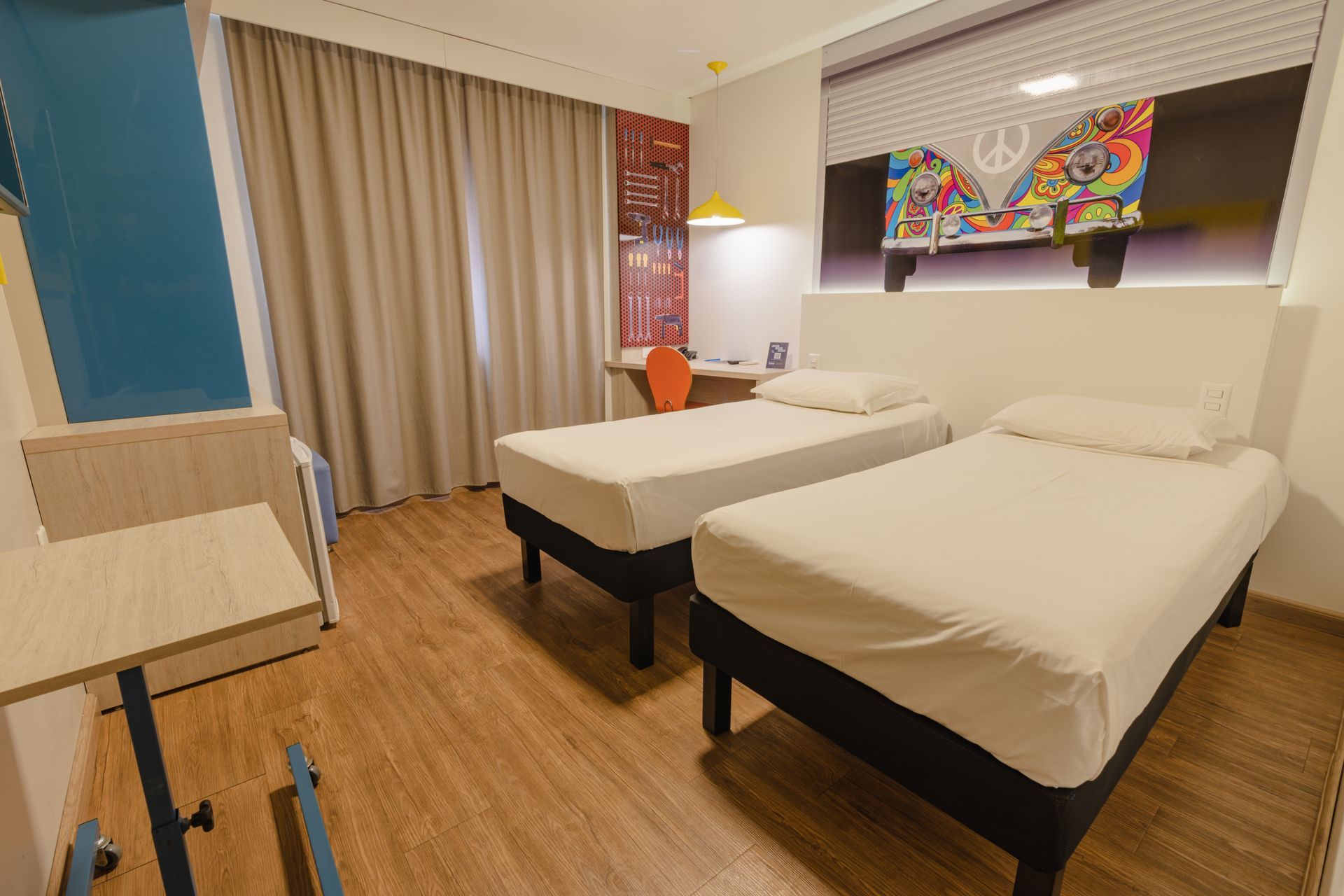 Quarto de hotel com duas camas, escrivaninha e TV na parede. Tons de creme, marrom e bege.