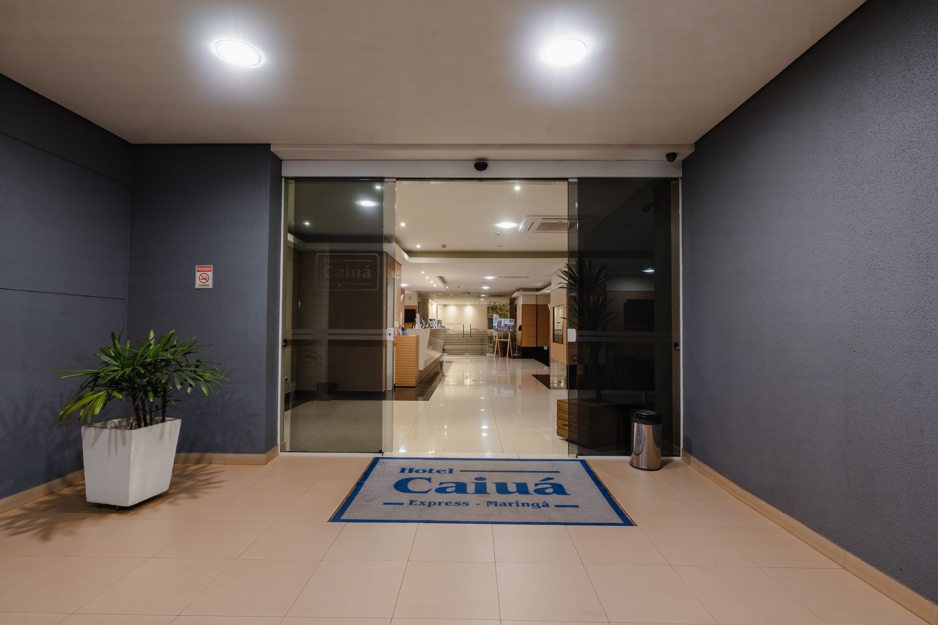 Hall de entrada com janelas do chão ao teto, bar, mesas de madeira e escadaria. Ambiente luminoso com piso de azulejo branco.
