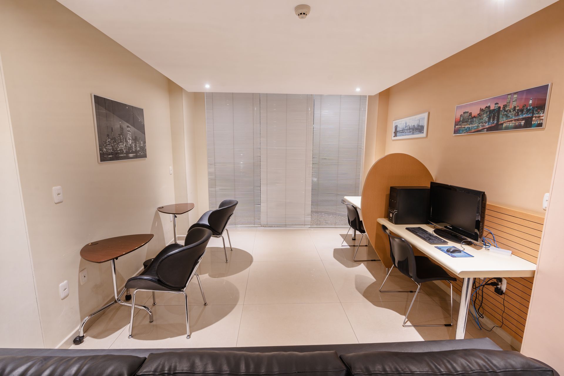 Quarto de hotel com cama, escrivaninha com TV, cadeira e sofá preto. Cortinas bege e piso de madeira.