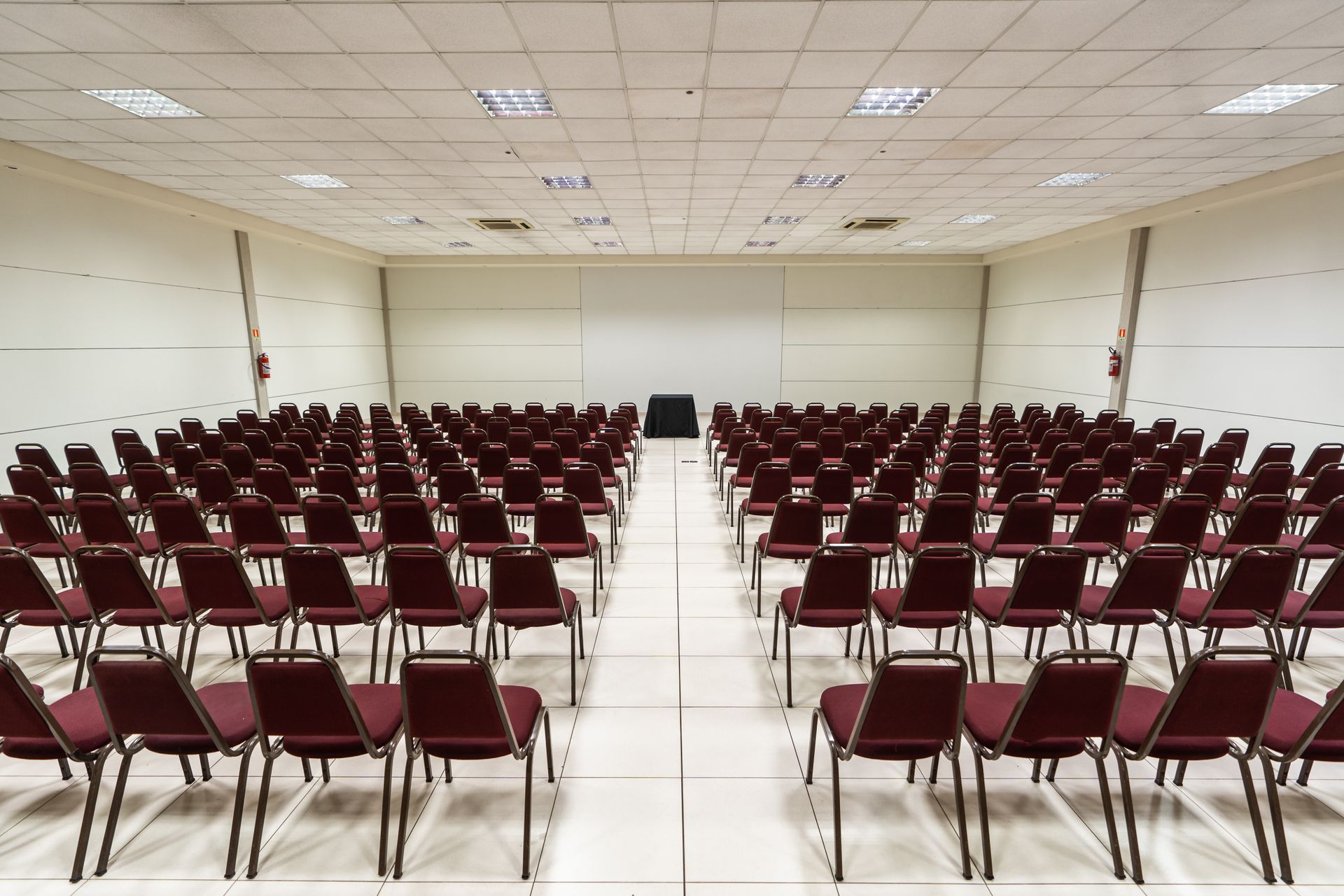 Sala de eventos com fileiras de mesas cobertas com toalhas, cadeiras azuis, paredes em tons neutros e iluminação no teto.
