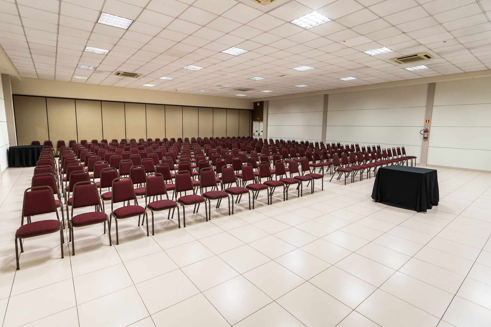 Sala de conferências vazia com fileiras de mesas e cadeiras pretas, paredes e teto brancos.