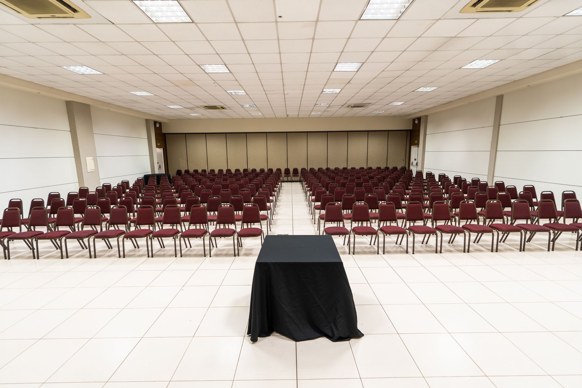 Sala de conferências vazia com fileiras de mesas cobertas com toalhas pretas.
