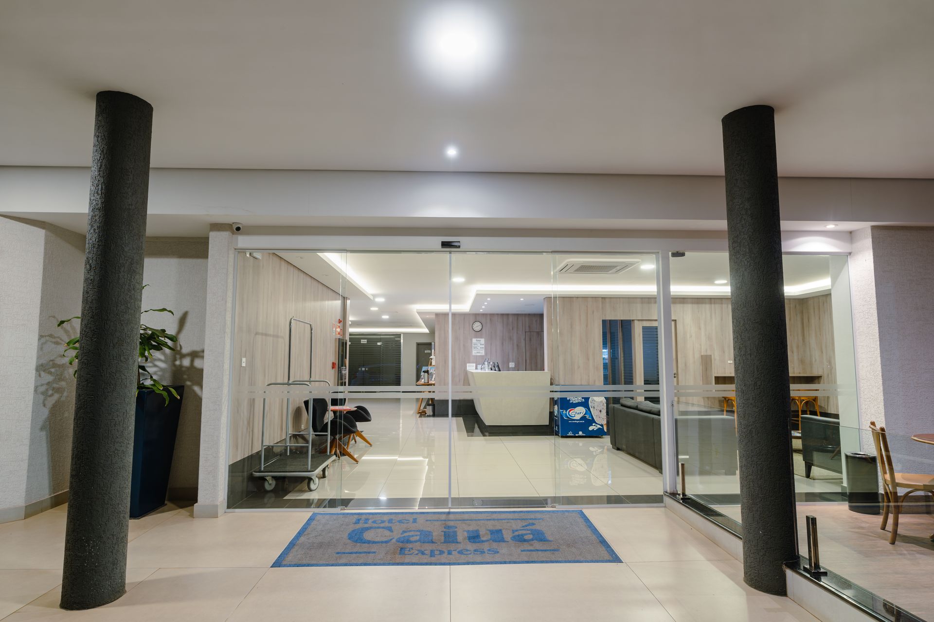Hall de entrada moderno com portas de vidro, bancos e duas colunas pretas sob luzes de teto brilhantes.