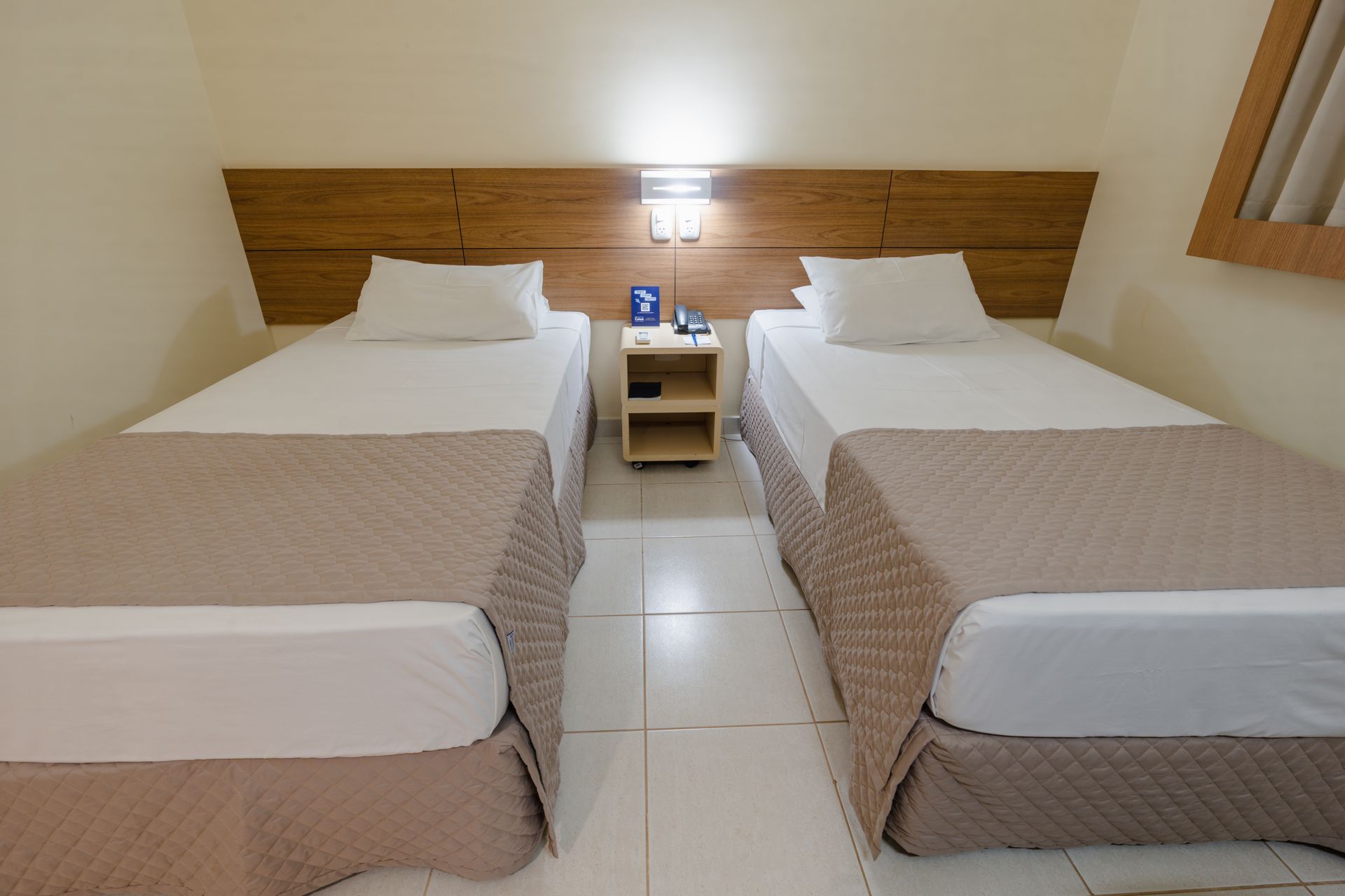 Quarto de hotel com duas camas, escrivaninha e TV na parede. Tons de creme, marrom e bege.