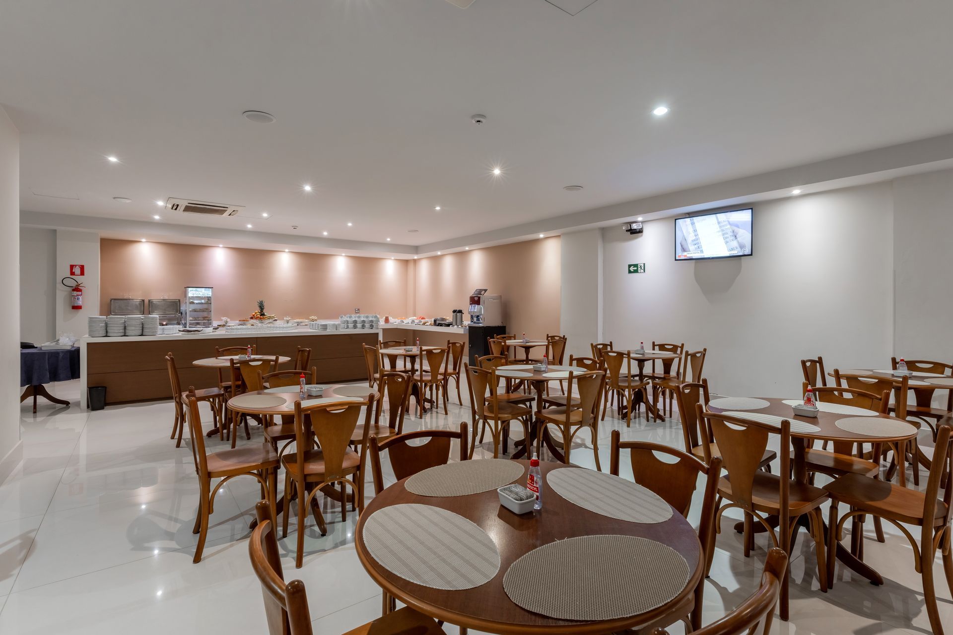 Sala de jantar do hotel com mesas redondas, cadeiras de madeira e um bufê com comidas e bebidas.