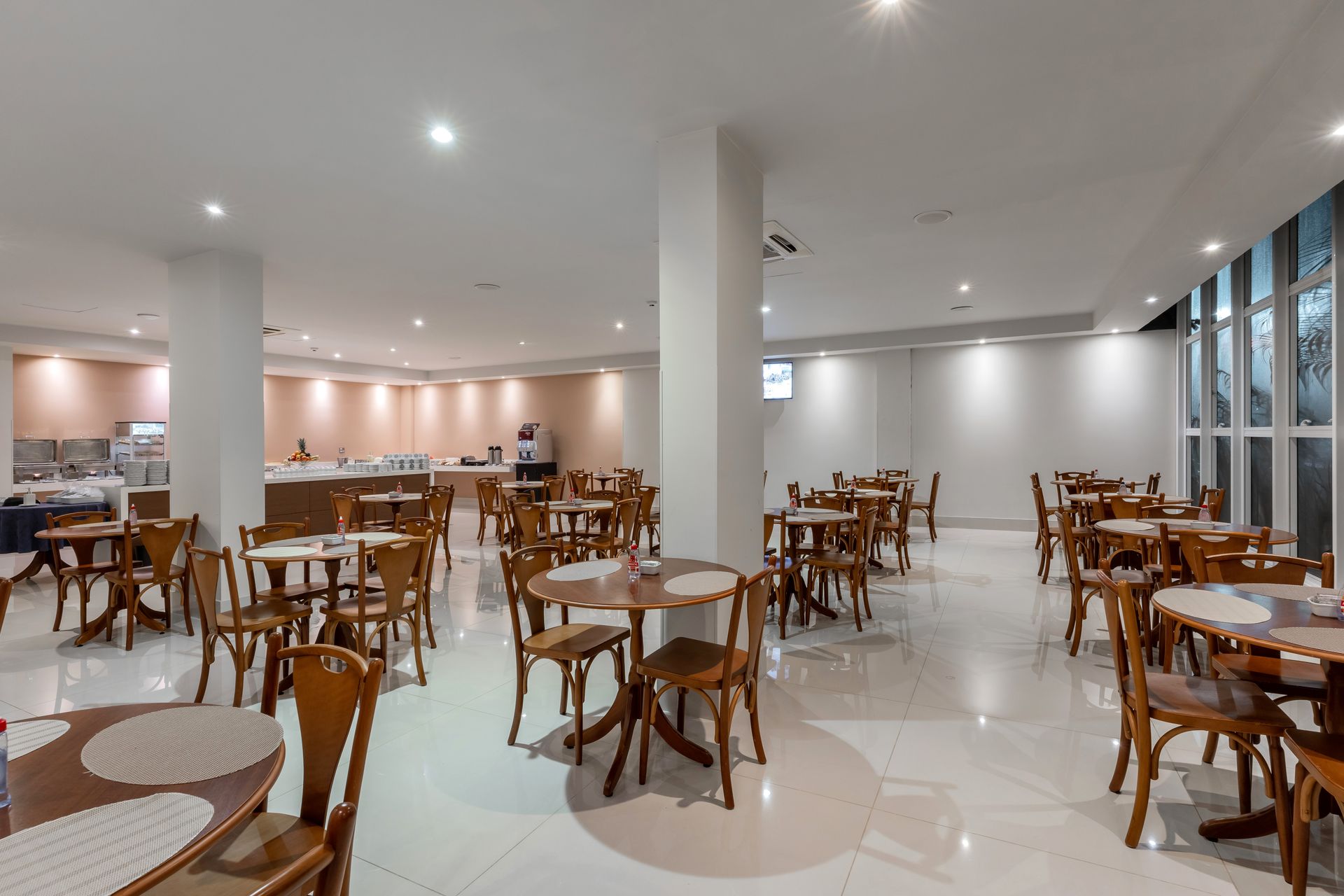 Sala de jantar com mesas e cadeiras redondas, e um buffet ao fundo. Paredes brancas, móveis de madeira e iluminação clara.