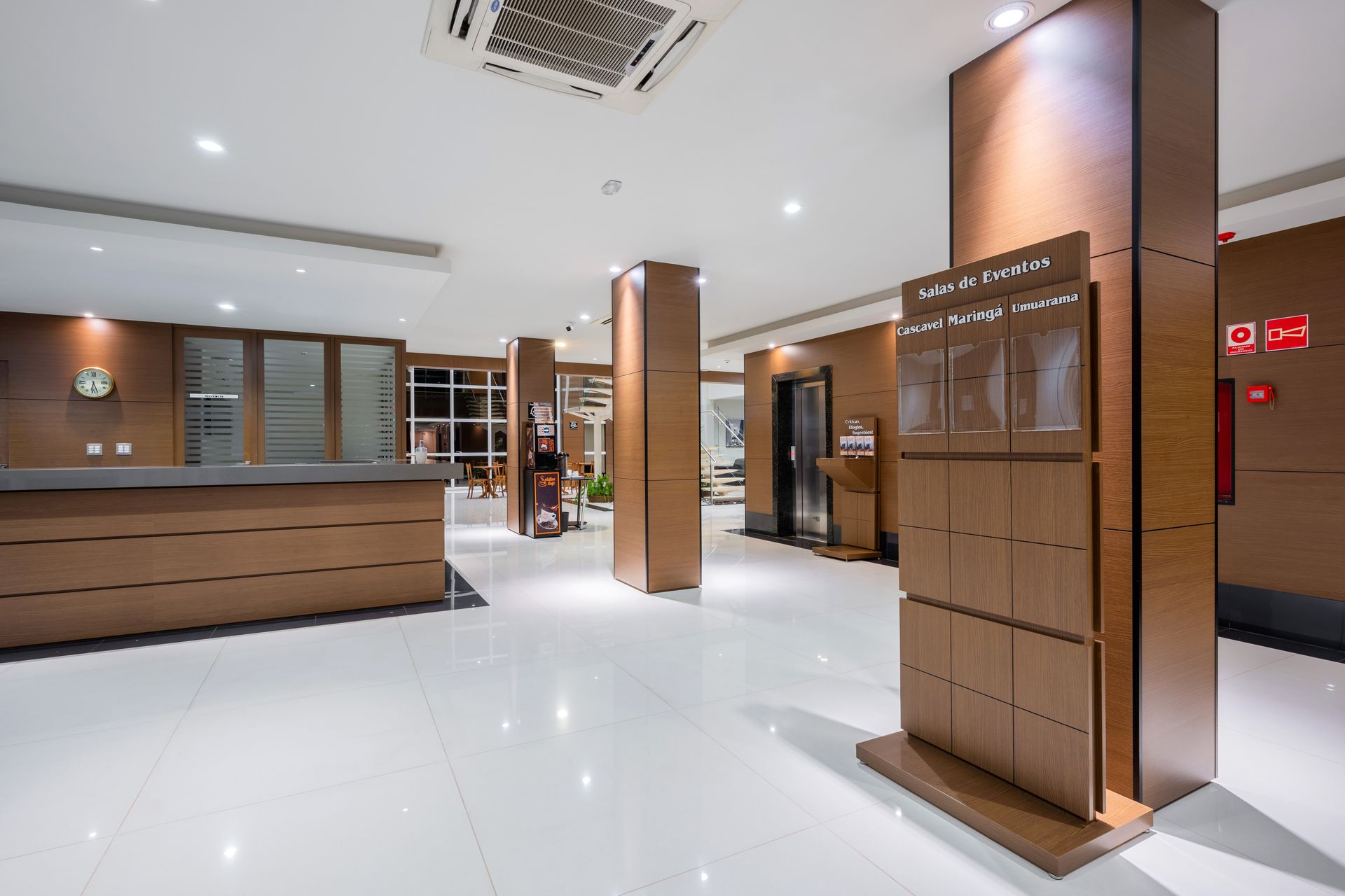 Hall de entrada do hotel com balcão de recepção em madeira marrom, pilares e piso branco brilhante.