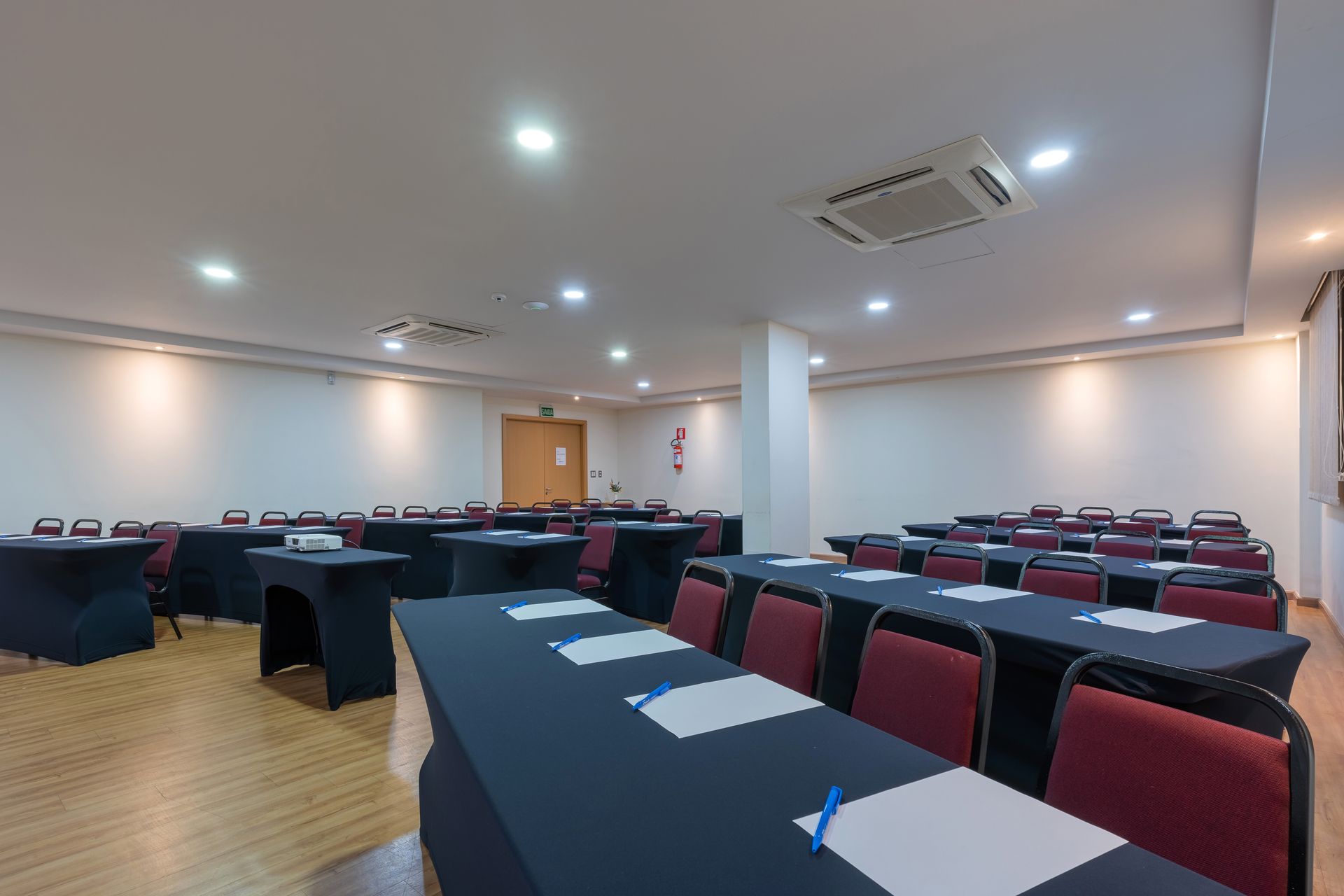 Sala de conferências com mesas em formato de U, toalhas de mesa azul-escuras, cadeiras cinza e iluminação embutida.