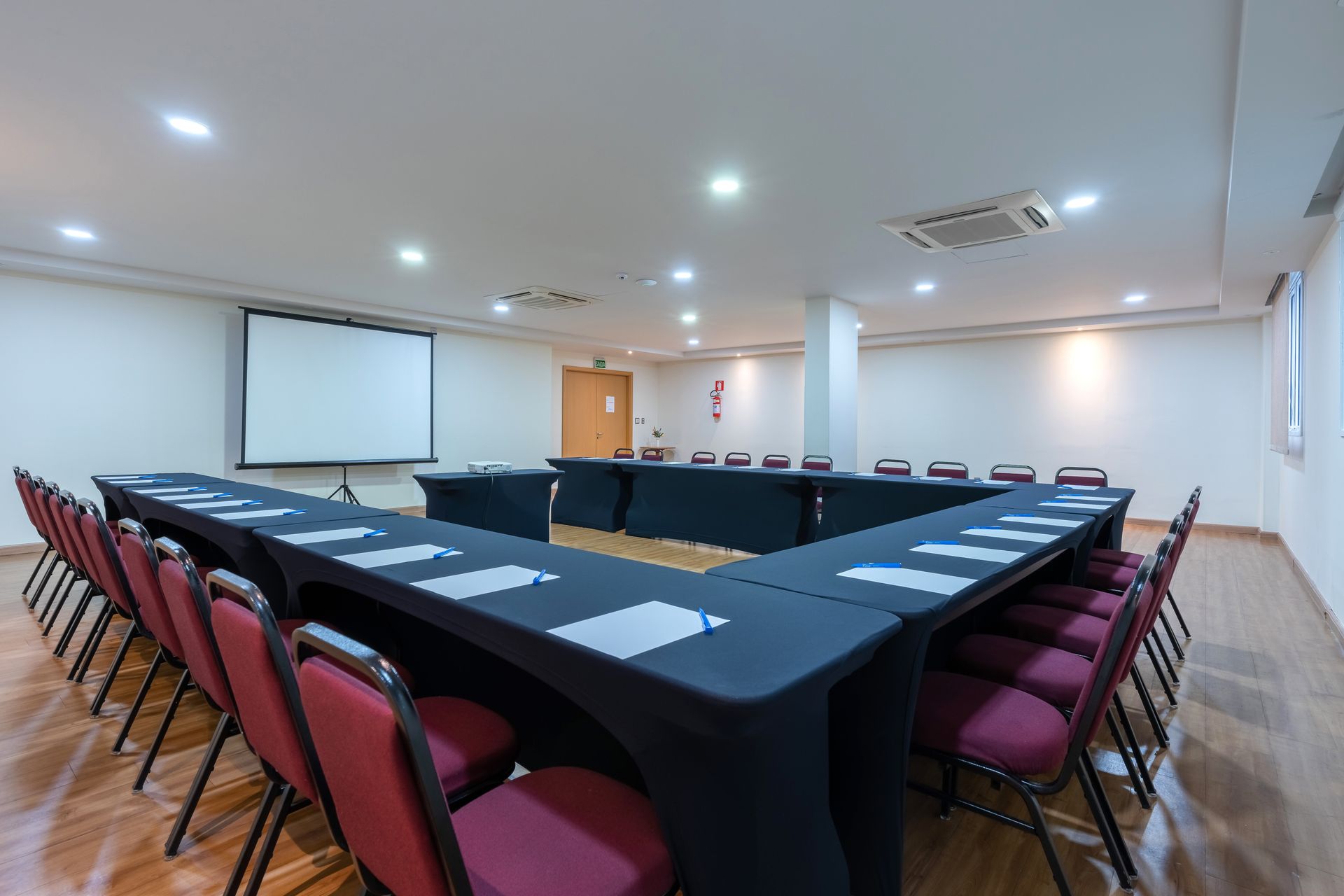 Sala de conferências com mesas dispostas em forma de U, cadeiras vermelhas, tela de projeção e piso de madeira.