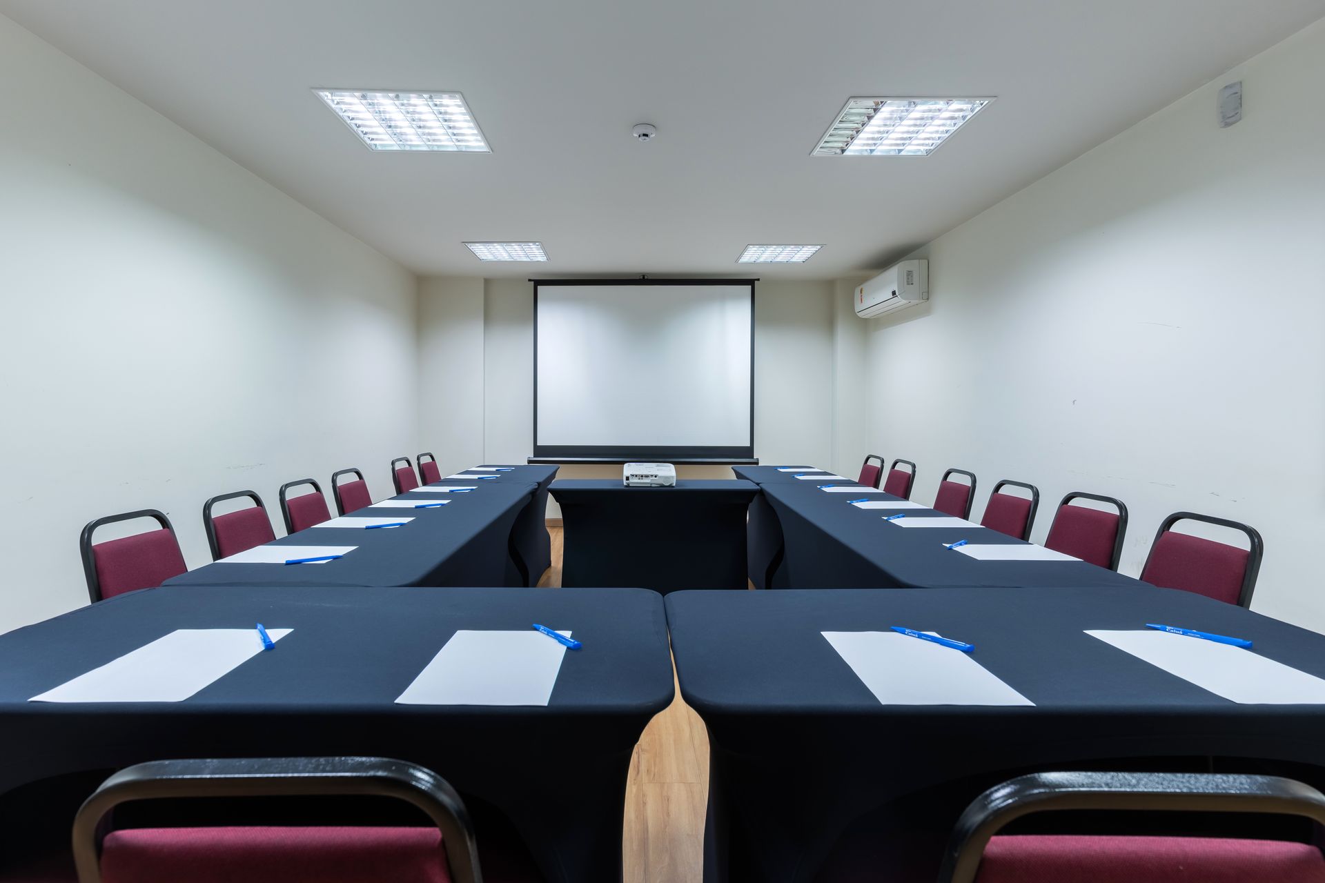 Sala de eventos com fileiras de mesas cobertas com toalhas, cadeiras azuis, paredes em tons neutros e iluminação no teto.