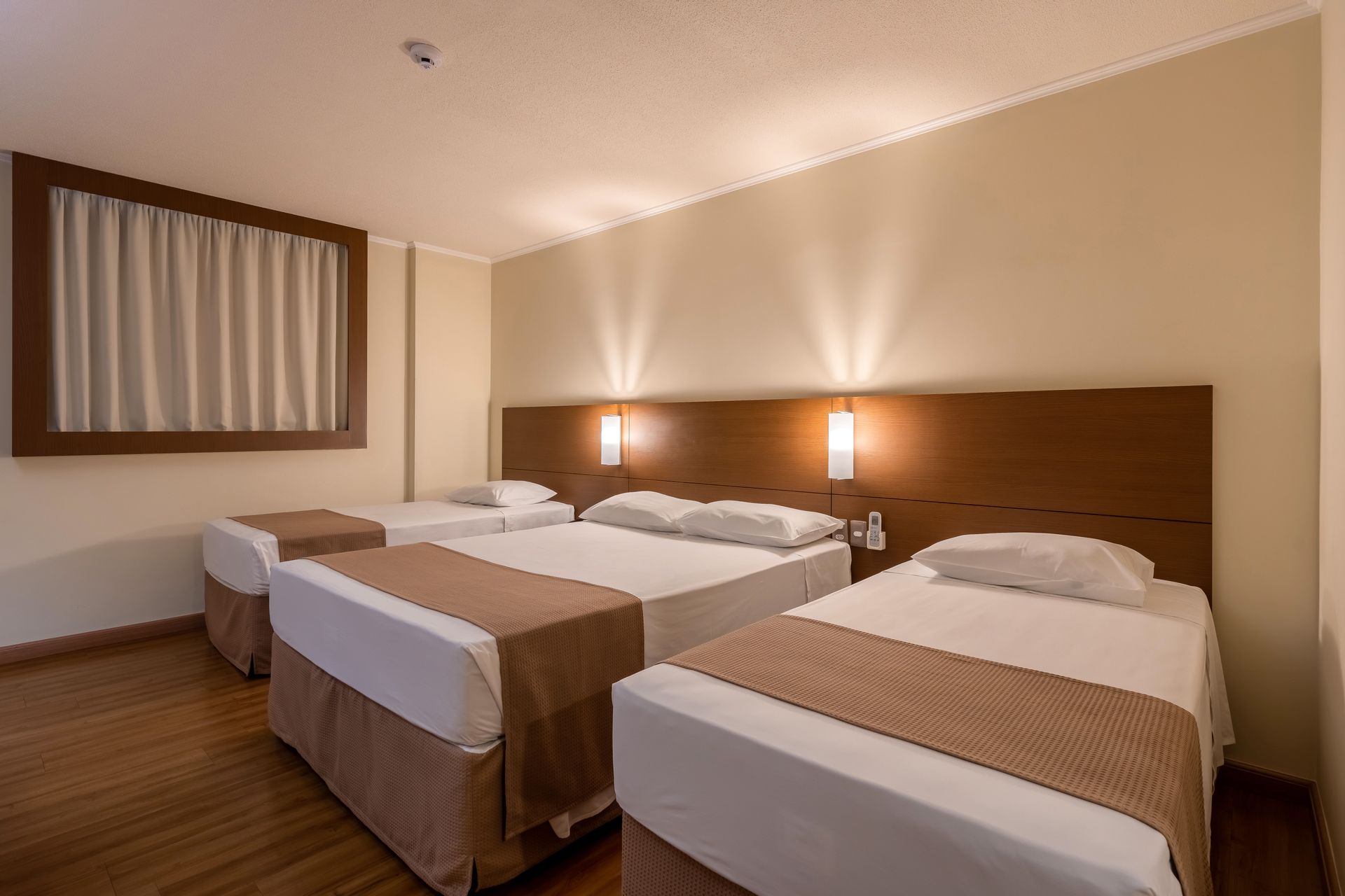 Quarto de hotel com três camas, janela com cortinas e iluminação aconchegante.