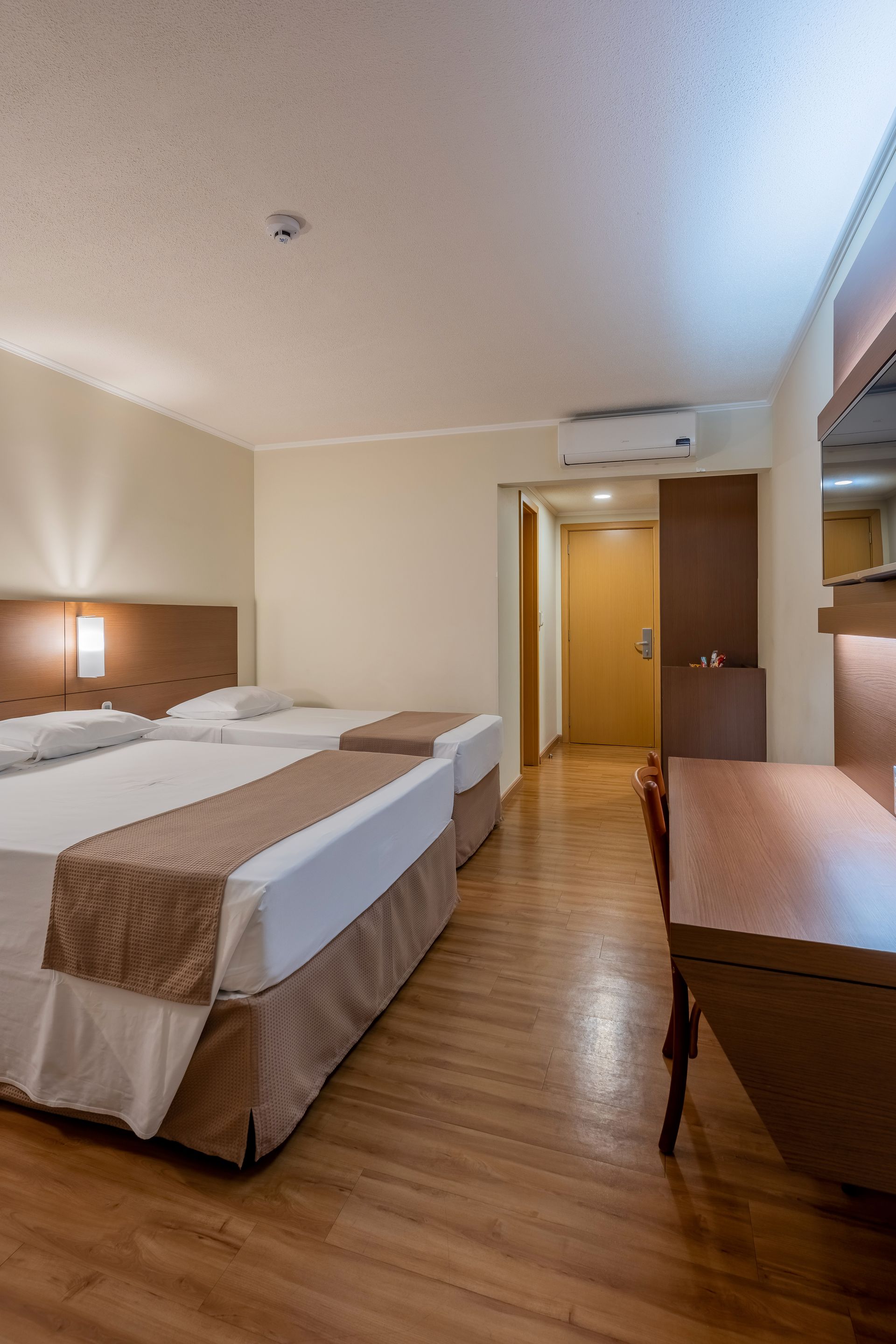 Quarto de hotel com três camas, uma escrivaninha e uma porta. Piso de madeira clara, paredes em tons neutros e peseiras bege.