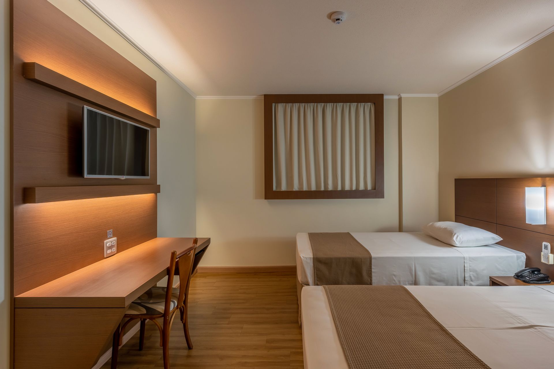 Quarto de hotel com duas camas de solteiro, escrivaninha e TV. Decoração em tons de marrom claro e bege.