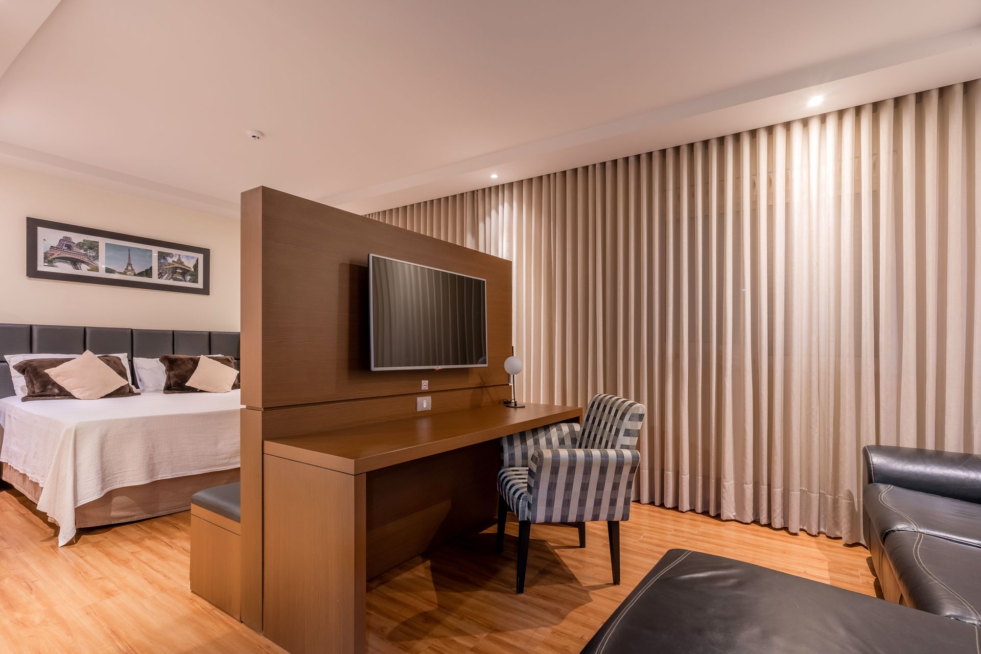Quarto de hotel com cama, escrivaninha com TV, cadeira e sofá preto. Cortinas bege e piso de madeira.