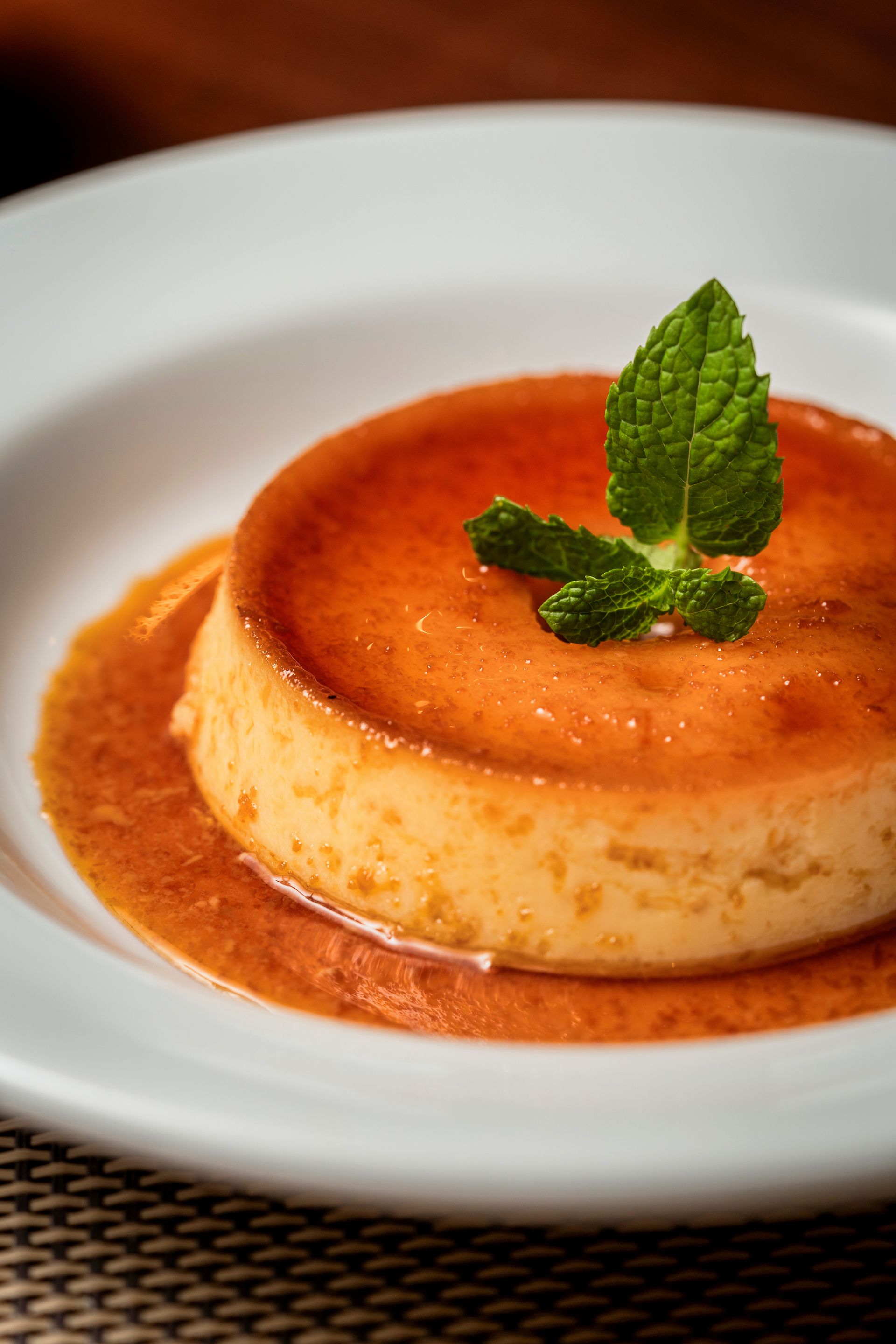 Sobremesa de pudim de leite com calda de caramelo, decorada com hortelã, servida em uma tigela branca.