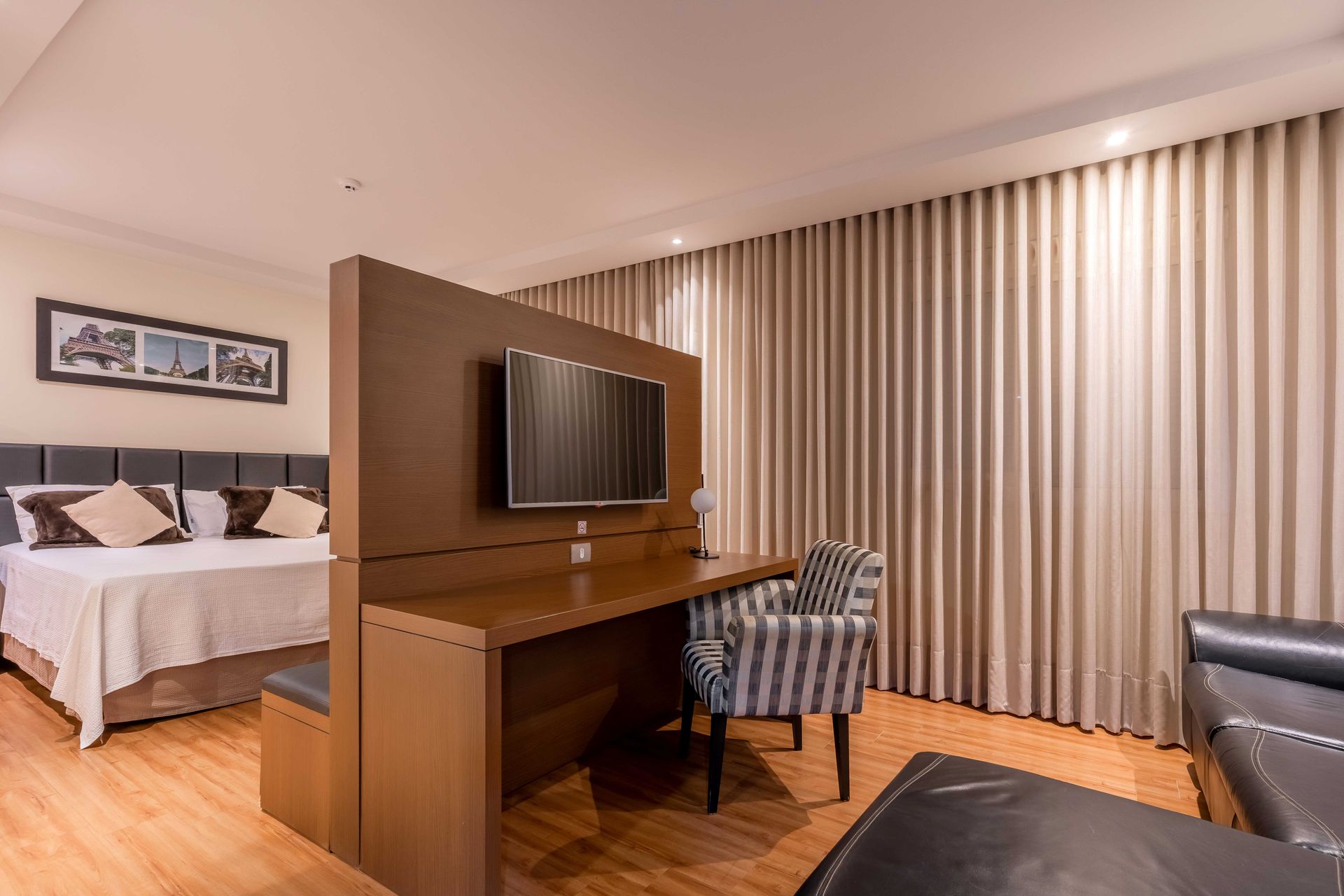 Quarto de hotel com cama, escrivaninha com TV, cadeira listrada e sofá. Tons de marrom e bege.