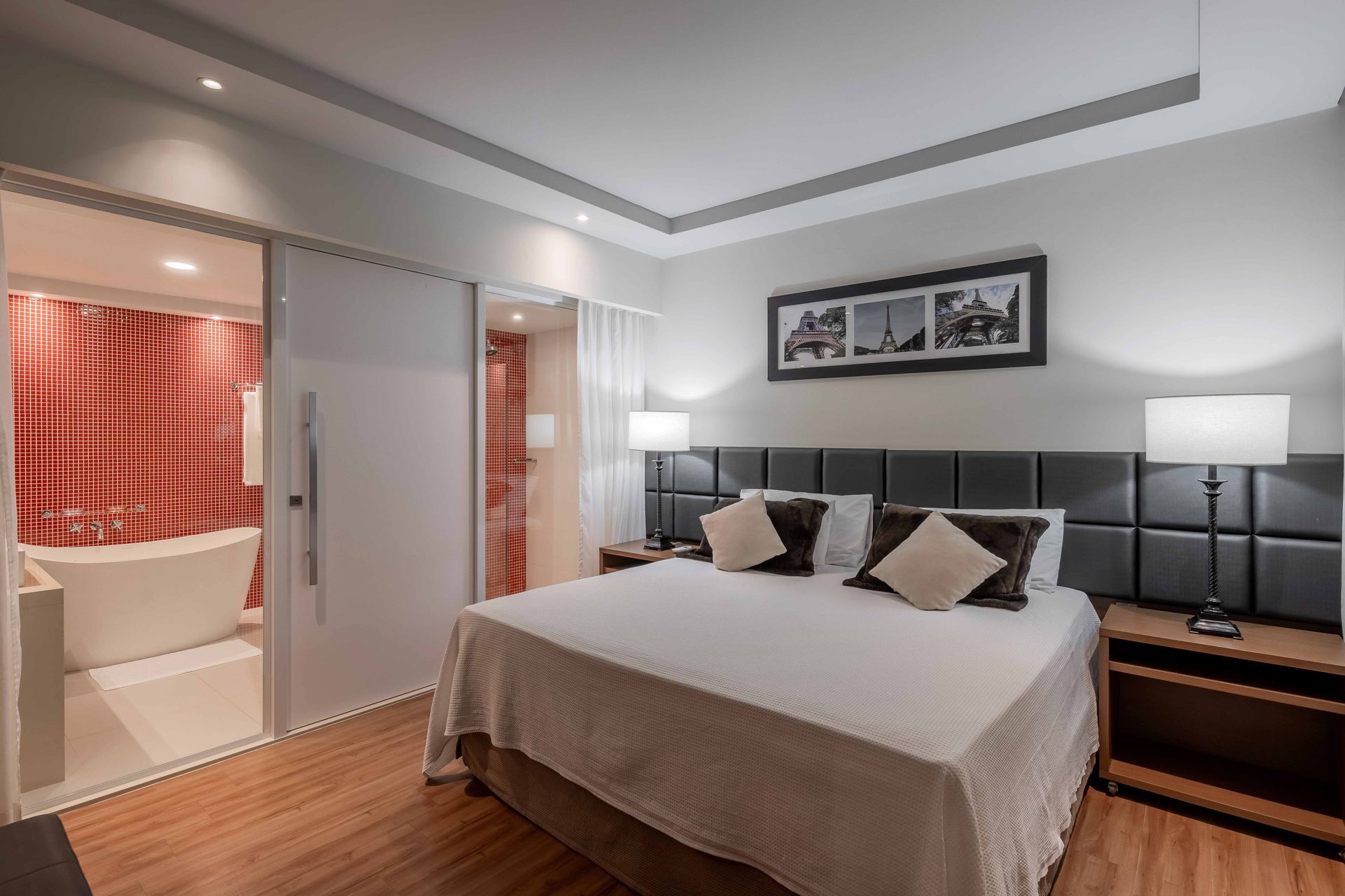 Quarto com cama, mesas de cabeceira e banheiro visível através de uma porta de vidro deslizante.