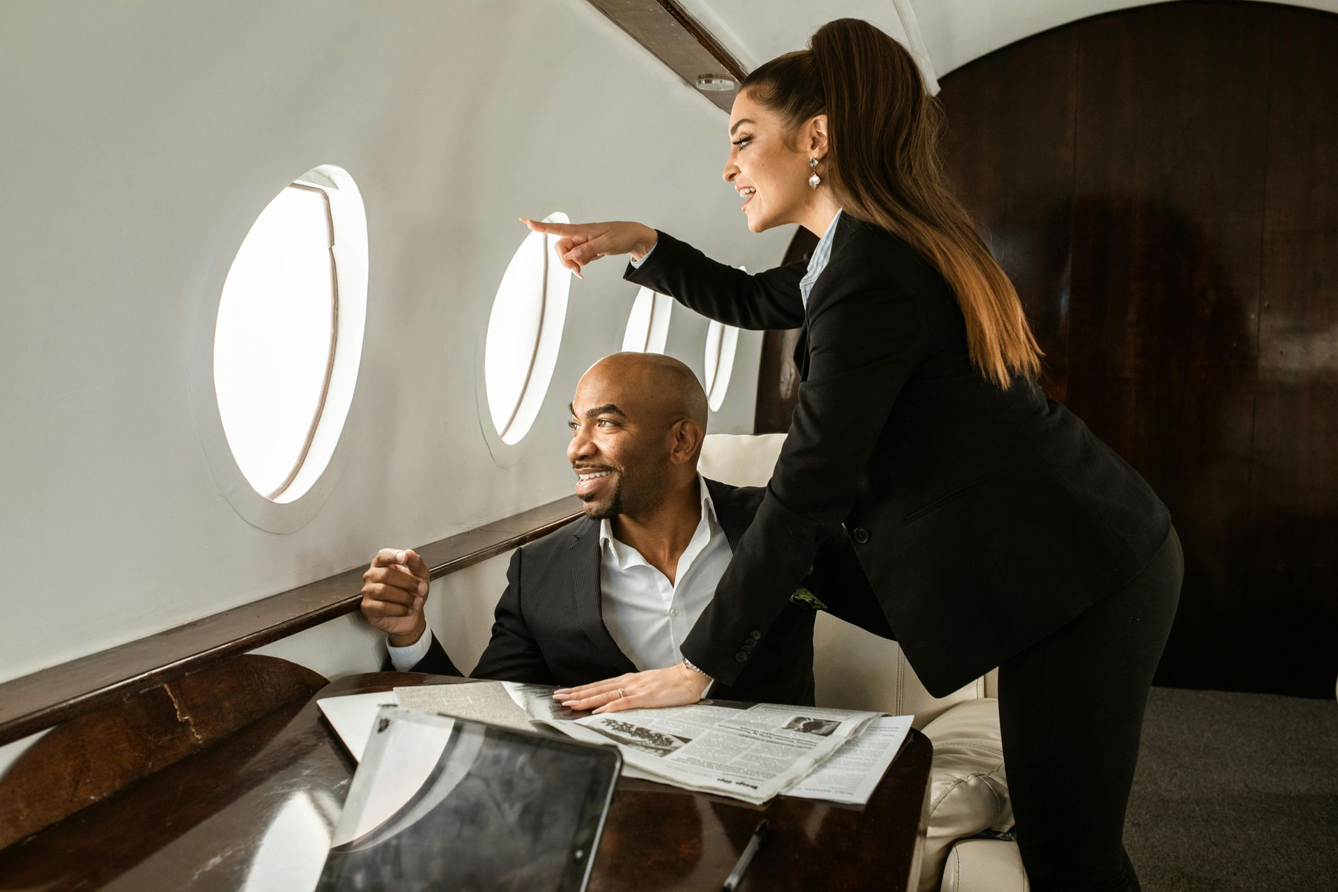 A businesswoman points out the window of a private jet to a Black man looking out, both dressed in suits.