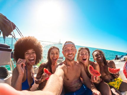 Group of friends on a boat selfie, holding watermelon, smiling, blue sky and ocean background.