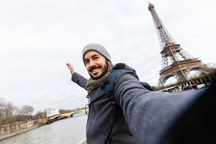 Man takes a selfie in Paris with the Eiffel Tower in the background, arms outstretched. Cloudy day.