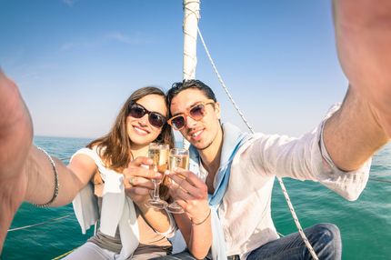 Couple on a sailboat taking a selfie, toasting champagne, blue water and sky.