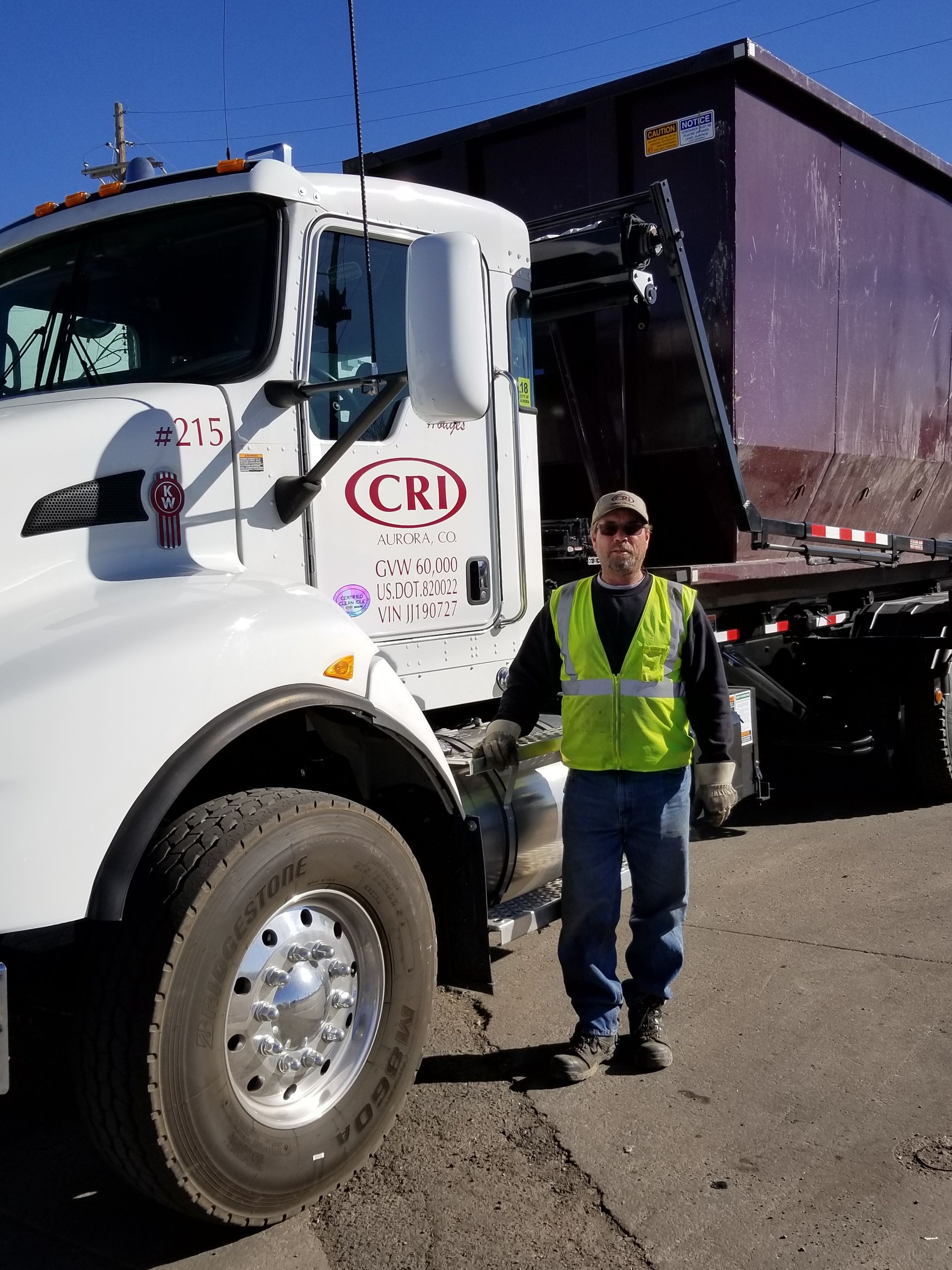 Driver in safety vest beside a roll-off truck hauling a dumpster rental