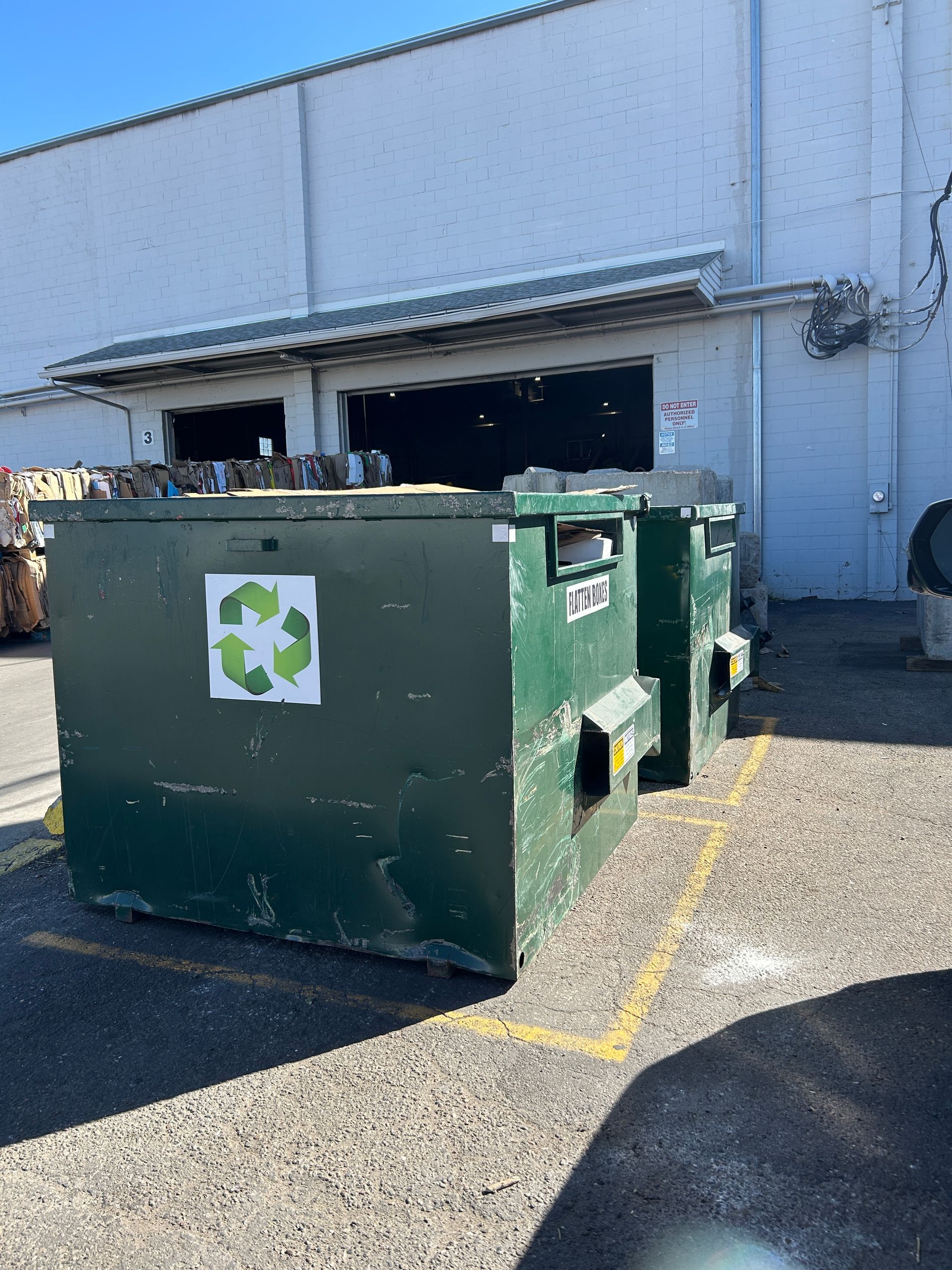 Green dumpsters with a recycling symbol beside a warehouse for roll-off dumpster rental