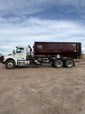 Photograph of a large white and maroon dump truck parked on a dirt surface under a partly cloudy sky