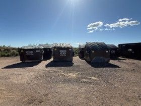Row of black front-load commercial trash dumpsters on a gravel lot under a clear blue sky