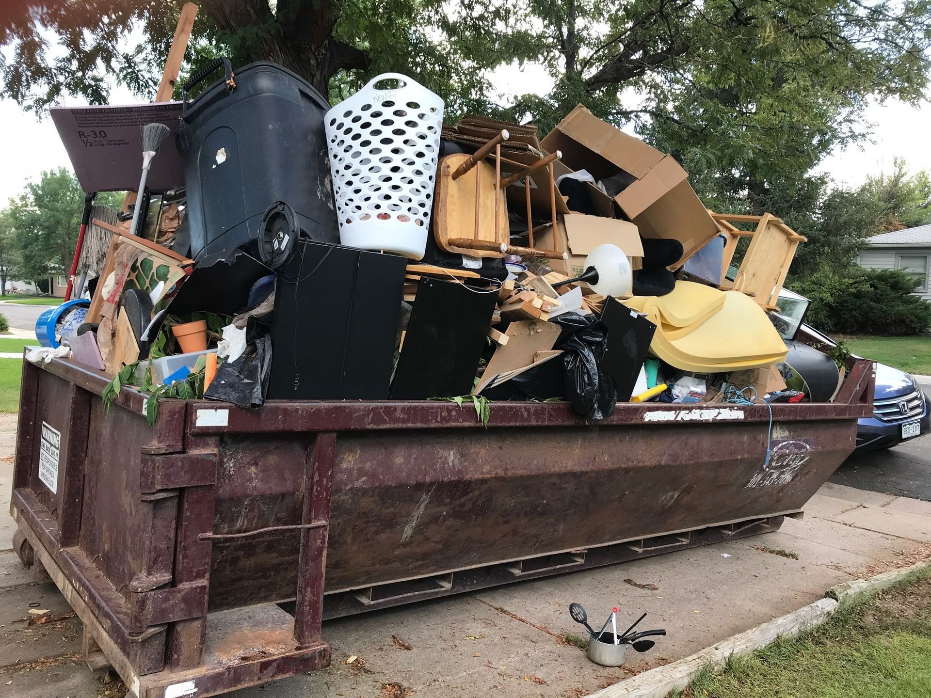 A dumpster overflowing with household junk sits on a concrete driveway near a street.