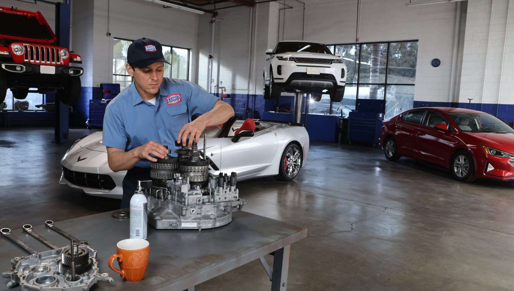 AAMCO mechanic working on a car transmission in a garage, with various vehicles in the background.