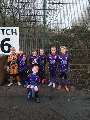 Youth soccer team in purple kits posing by a pitch sign on a wet field.