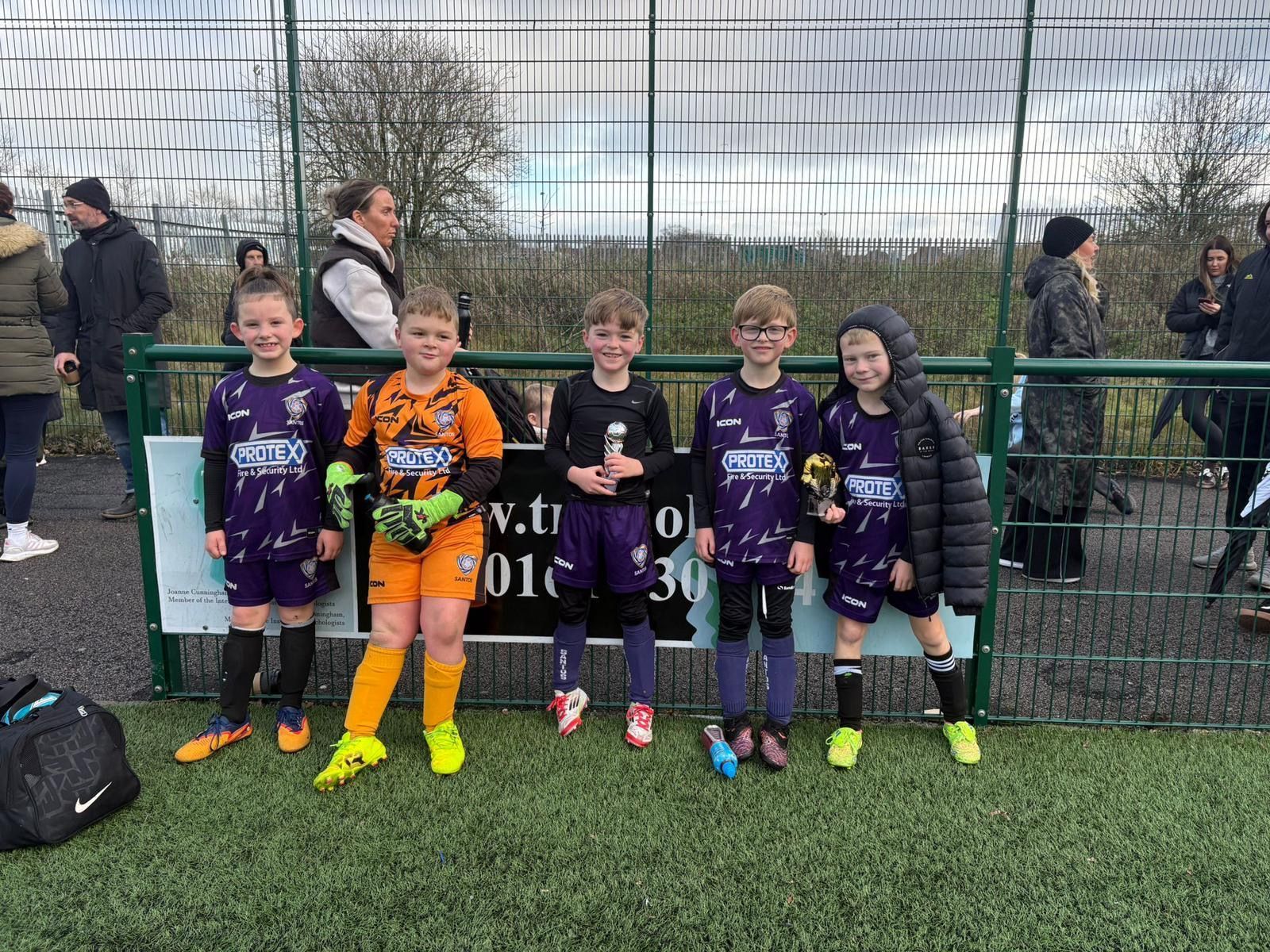 Five youth soccer players in black and purple uniforms posing outdoors by a fence with medals and a trophy