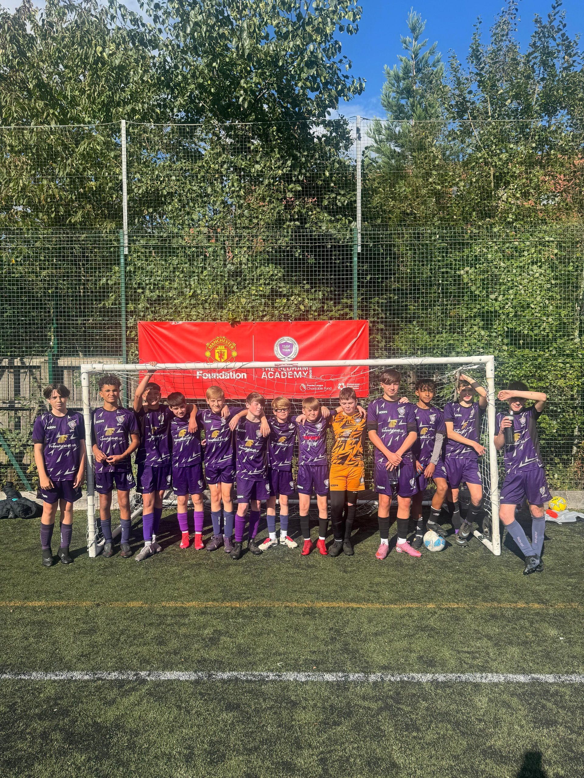 Youth soccer team posing in front of a goal on a sunny field, with a red banner behind them.